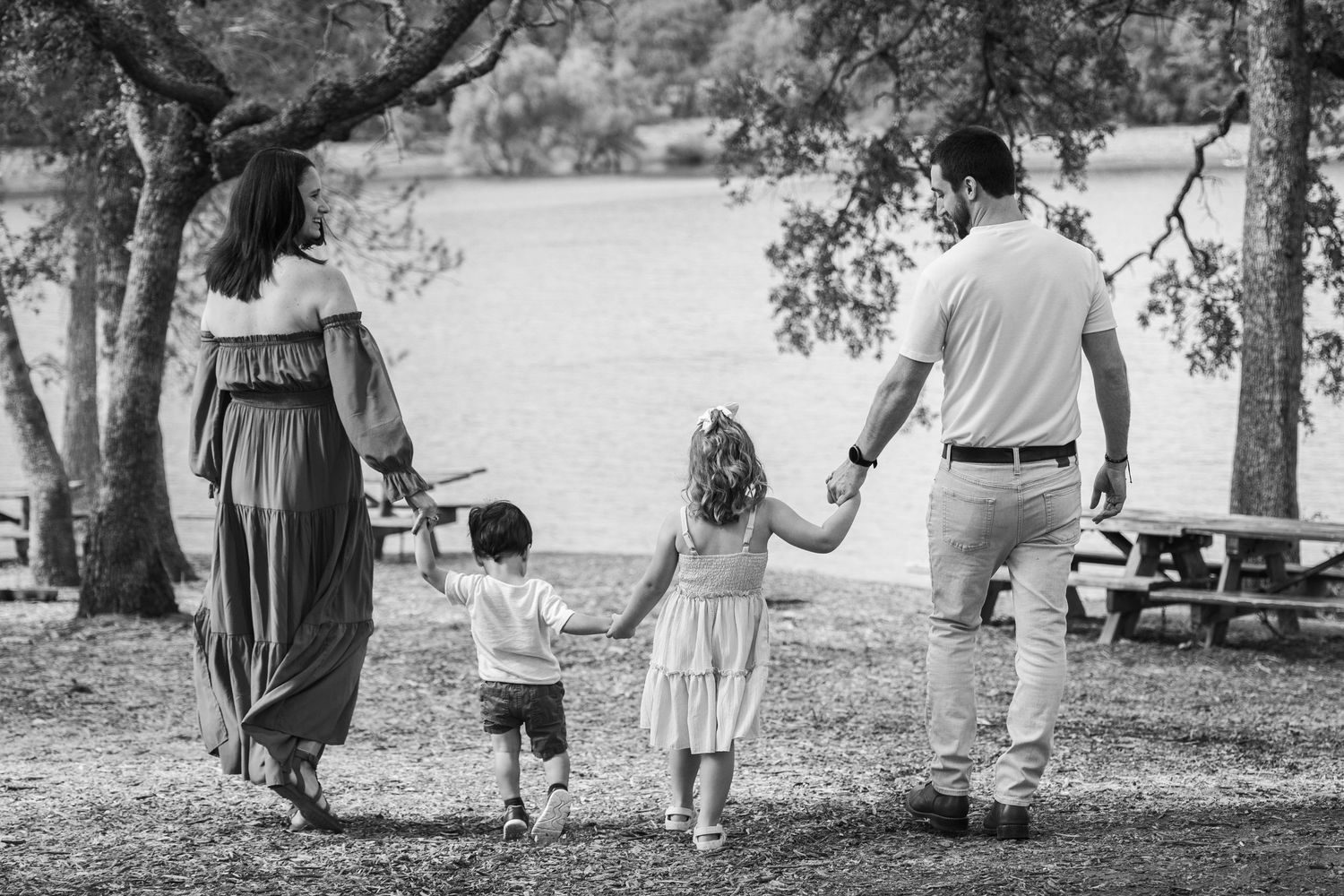 Black and white photo of a family of four holding hands walking toward a lake in a park.