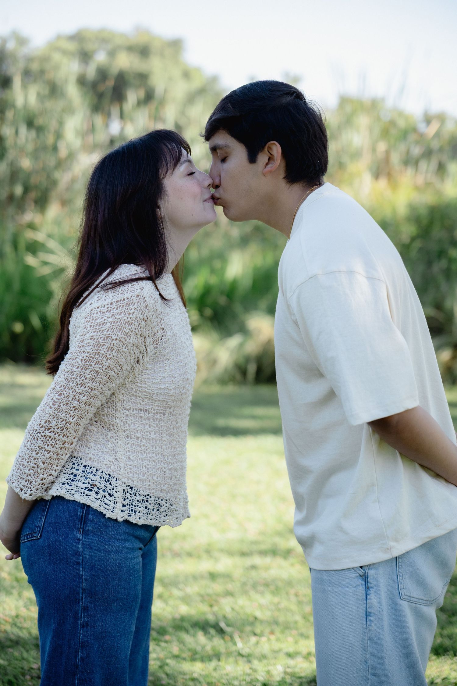 A young couple sharing a romantic kiss outdoors in a lush green park setting.