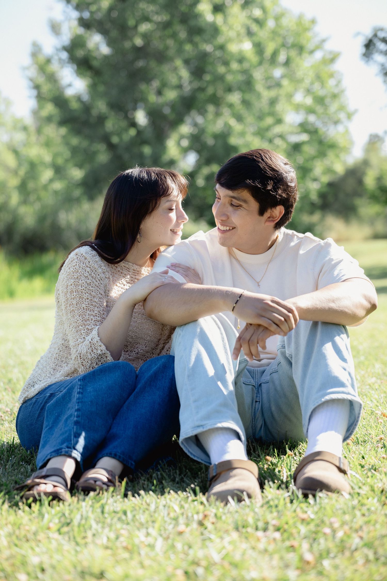 Young couple sitting on green grass in a sunny park, smiling and looking at each other affectionately.