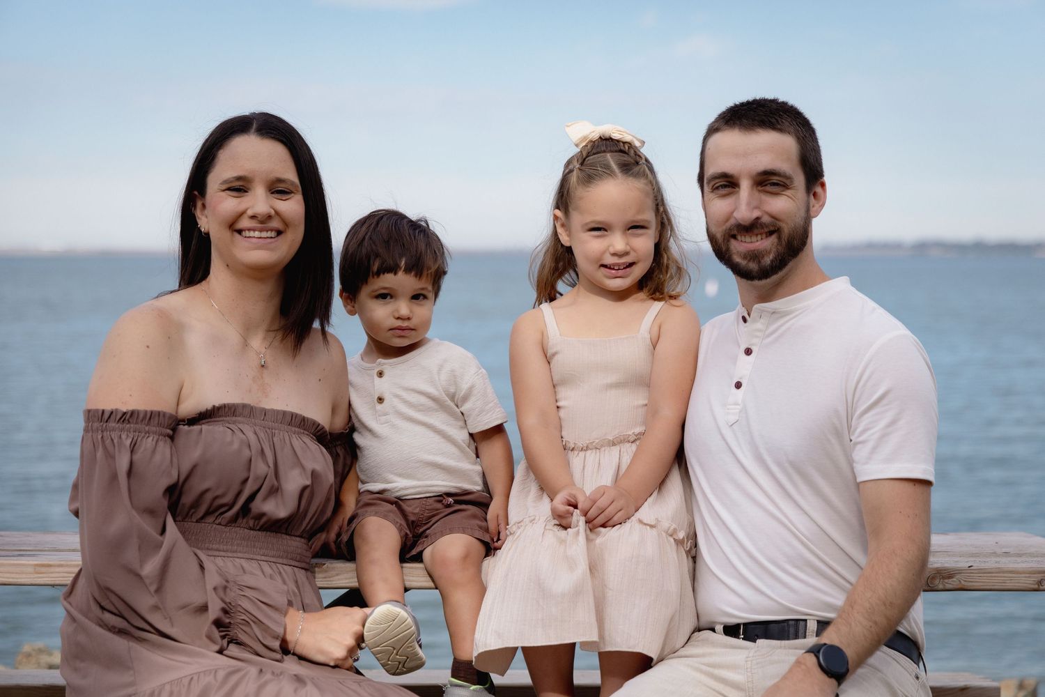 Family of four smiling together on a waterfront pier on a sunny day, dressed in coordinating neutral outfits.