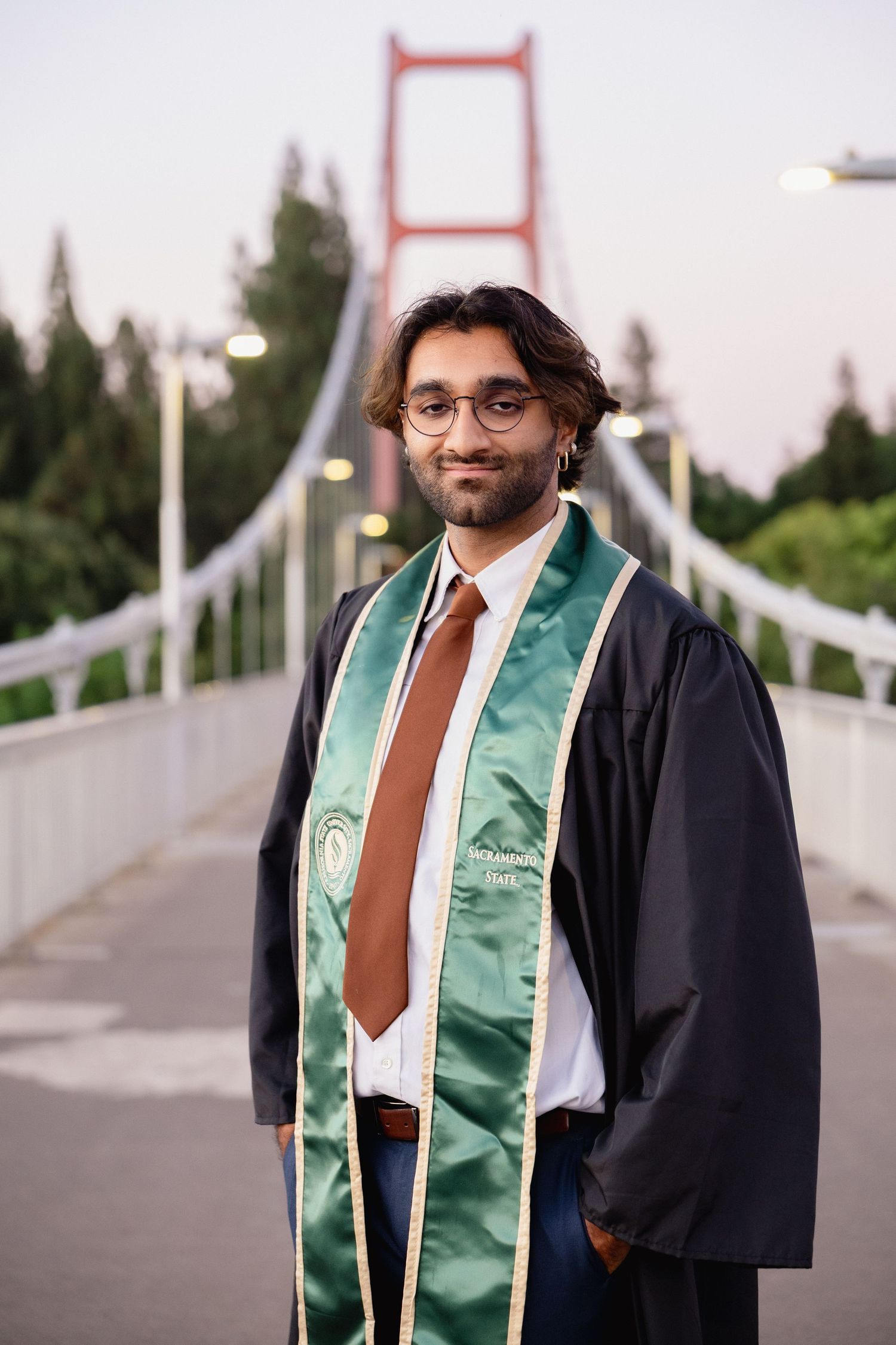 Graduate in black gown and green stole standing on bridge at dusk.
