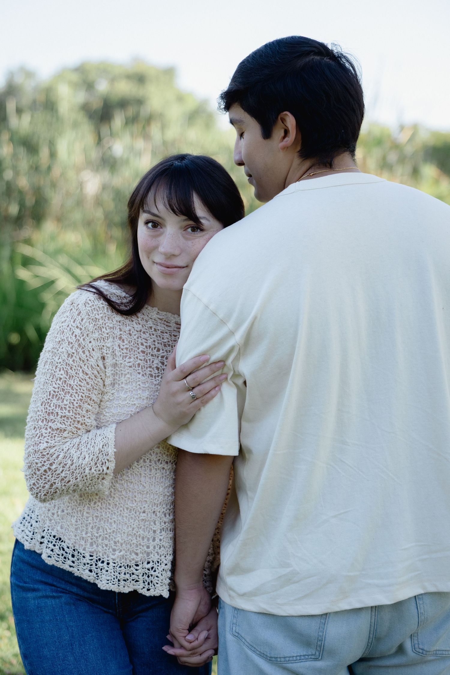 A young couple holds hands and shares a tender moment outdoors, surrounded by lush green trees.