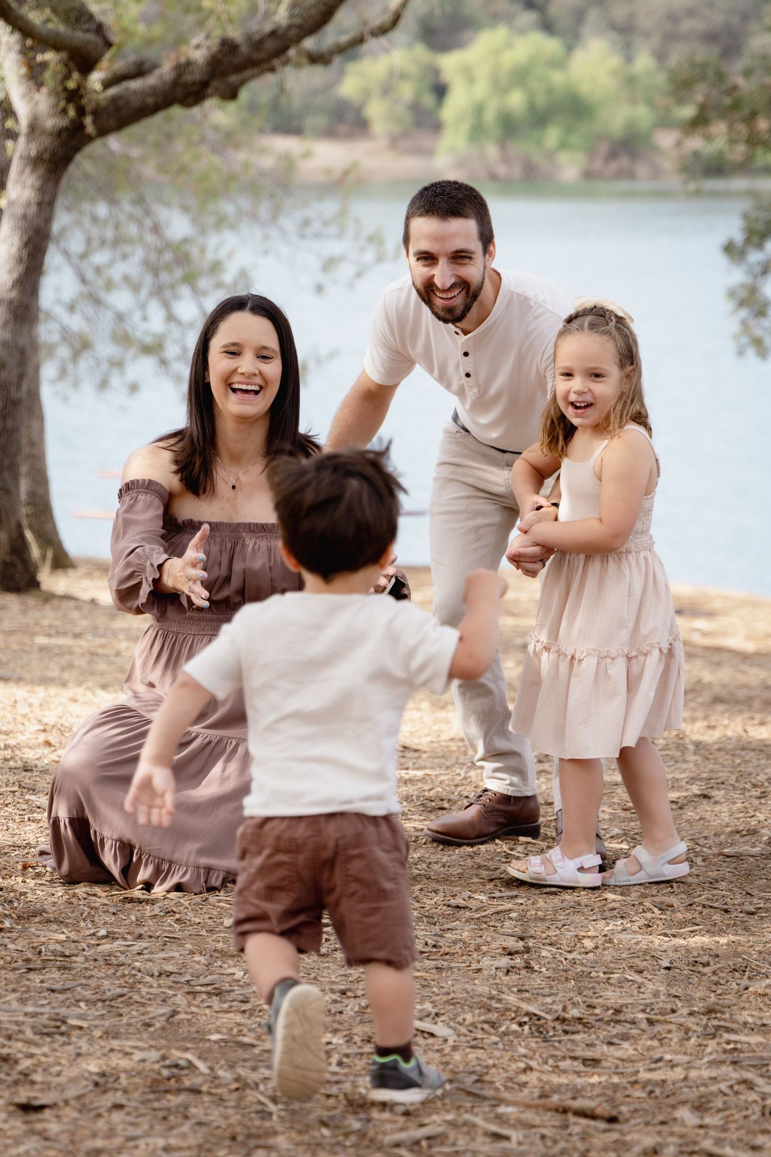 Happy family of four enjoying a sunny day at the park near a lake, with toddler running to parents.