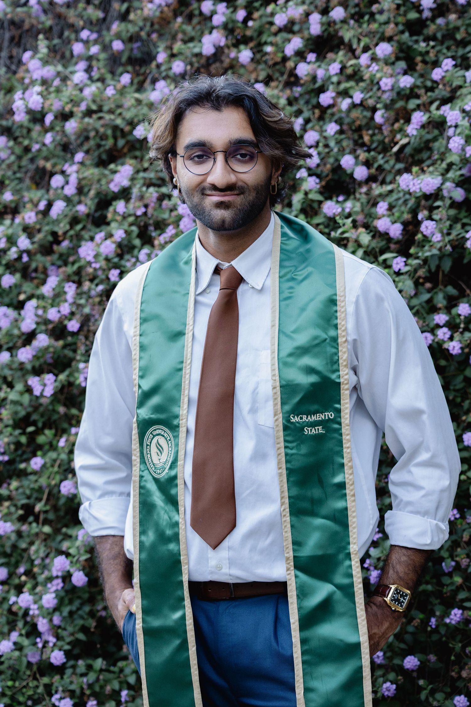 Sacramento State graduate smiling in green graduation stole, white shirt, and brown tie, standing before purple flowering bushes.