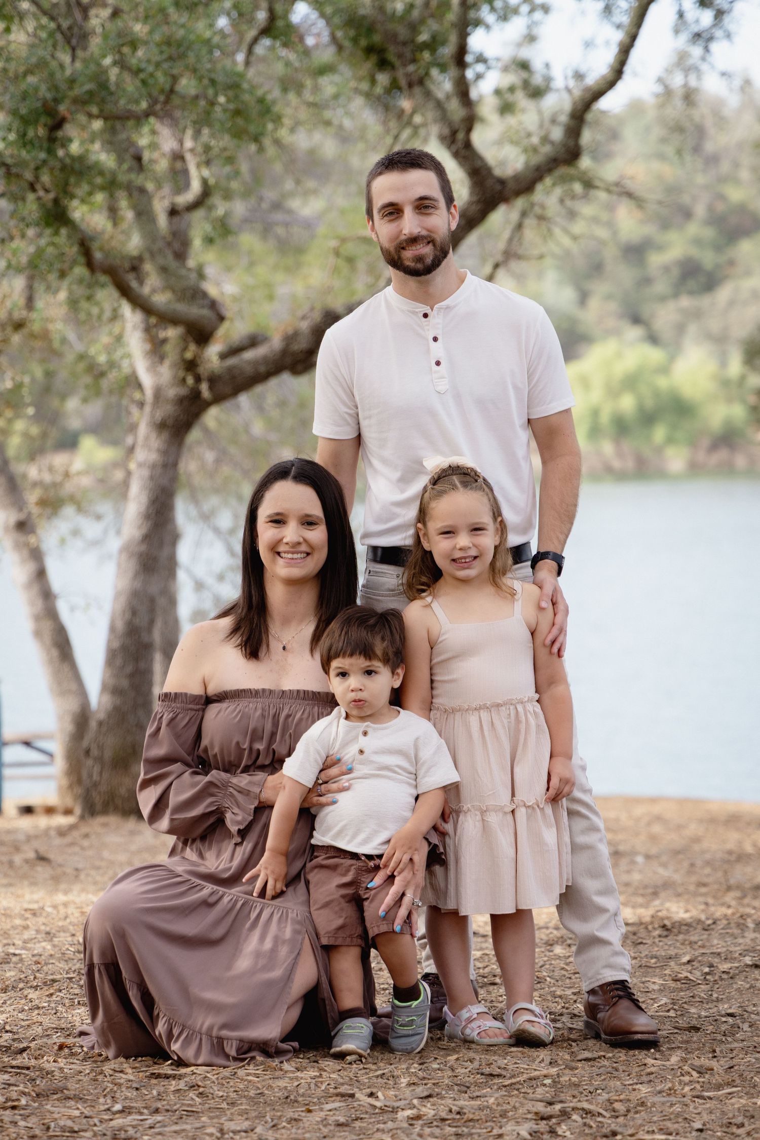 Family of four posing outdoors near a lake, with trees in background, parents and two young children smiling.