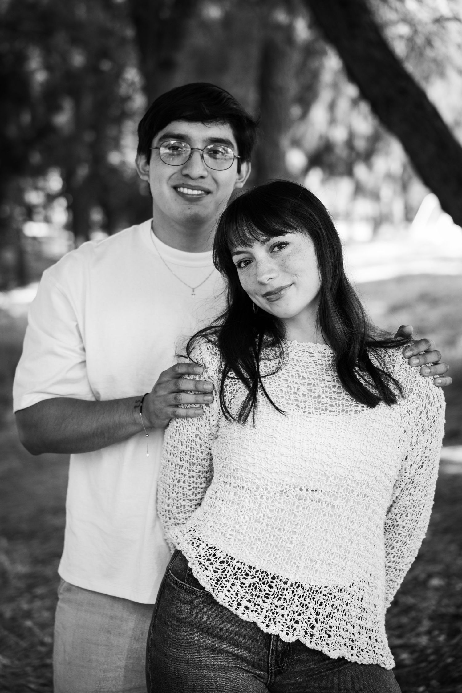 Black and white portrait of a young couple posing outdoors in a park, man in glasses with arm around woman.