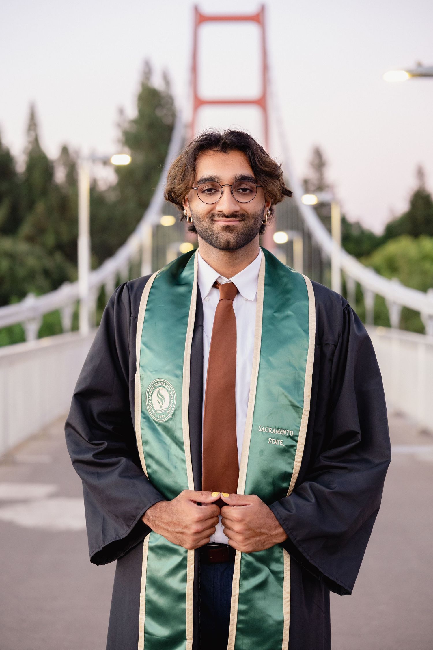 Graduate in black robe and green Sacramento State stole smiles on suspension bridge at dusk.