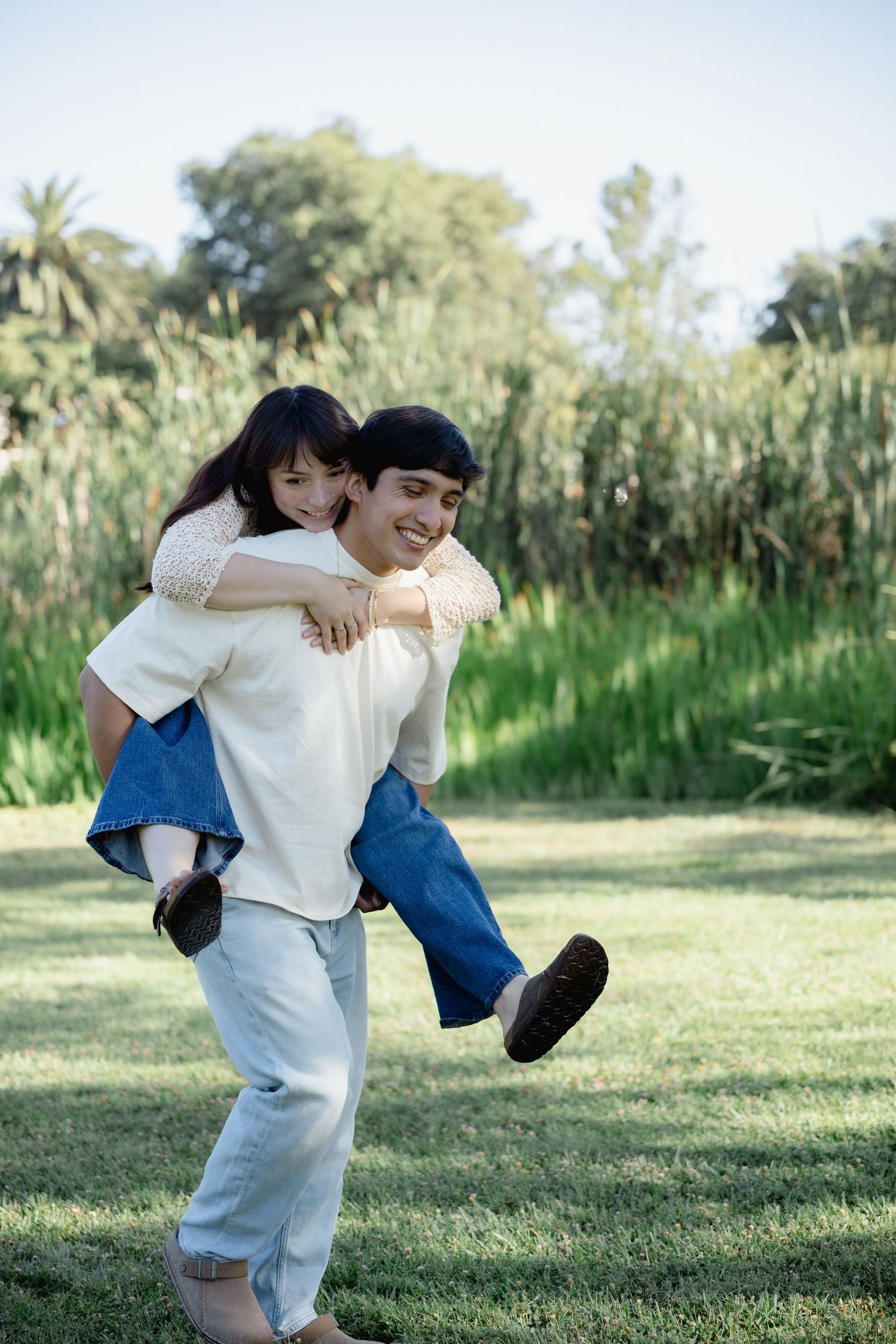 A smiling man gives a happy young woman a piggyback ride on a green lawn with trees in the background.