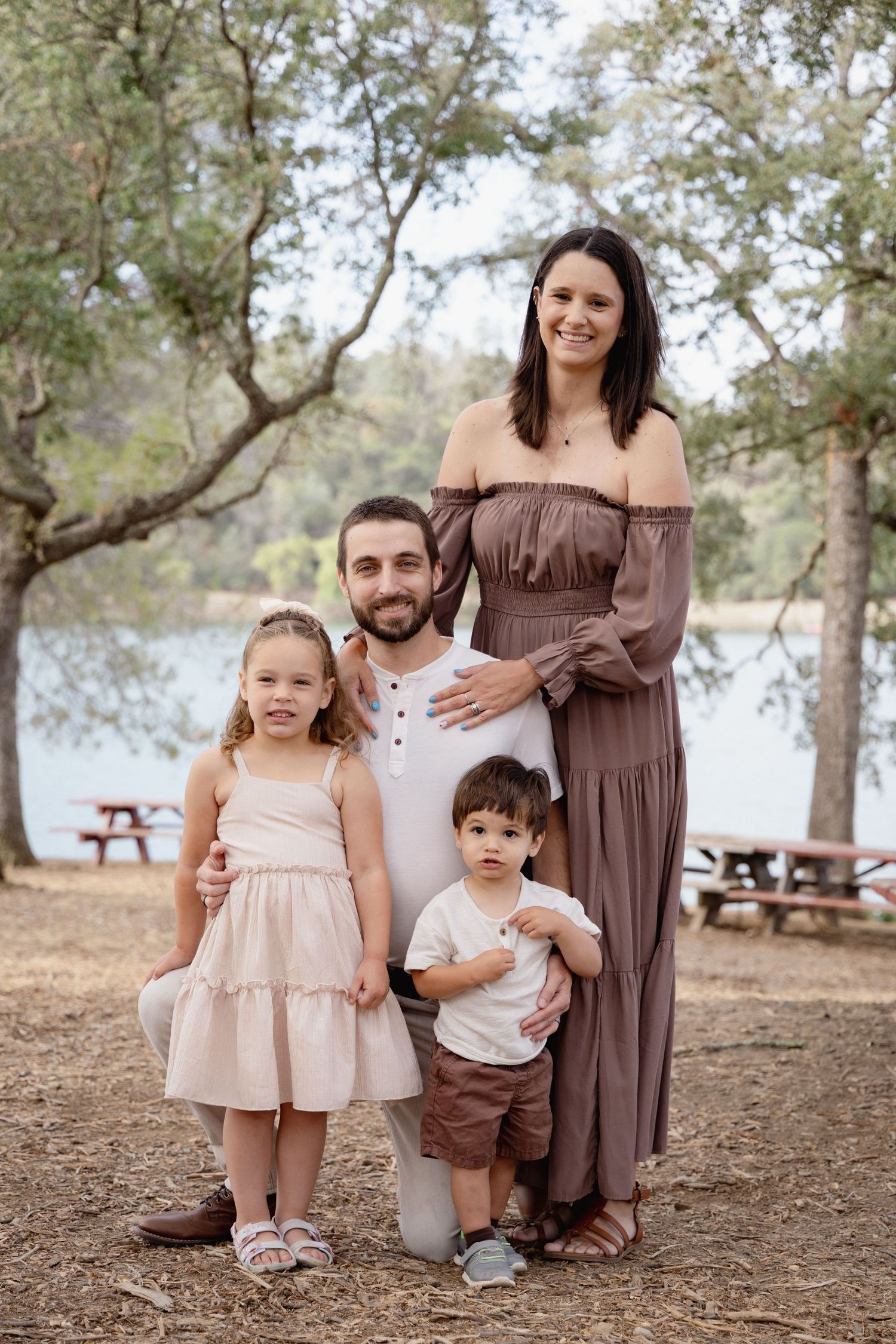 A smiling pregnant woman and bearded man pose outdoors with two young children near a lake surrounded by trees.