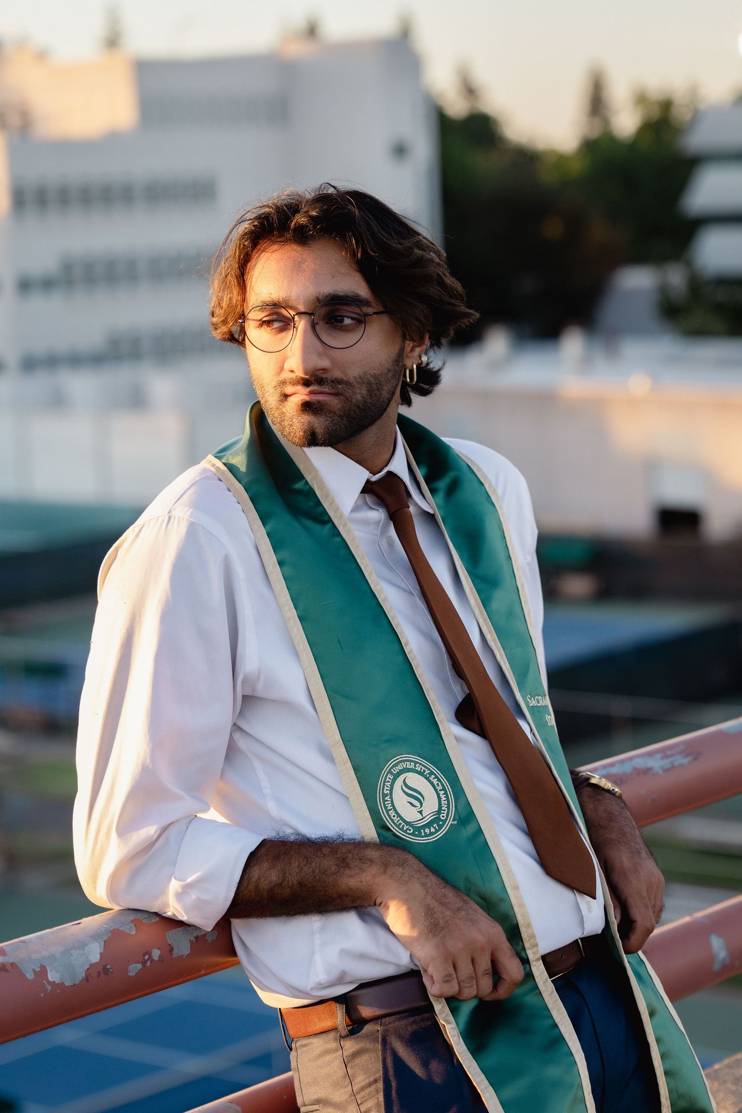 Young man in graduation sash and glasses poses on rooftop in white shirt and brown tie at sunset.