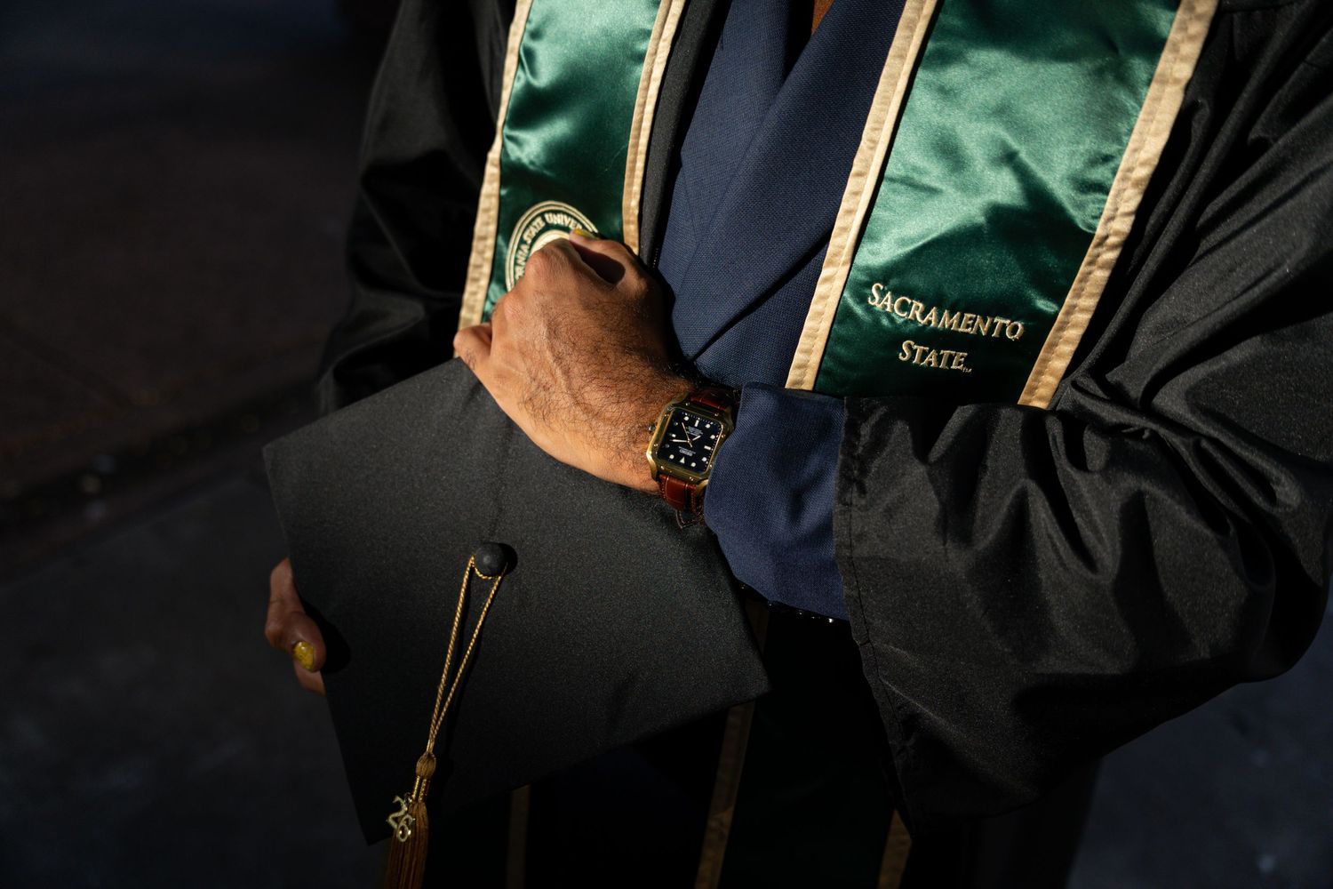Close-up of a Sacramento State graduation stole worn over a suit, with a mortarboard cap held in hand.