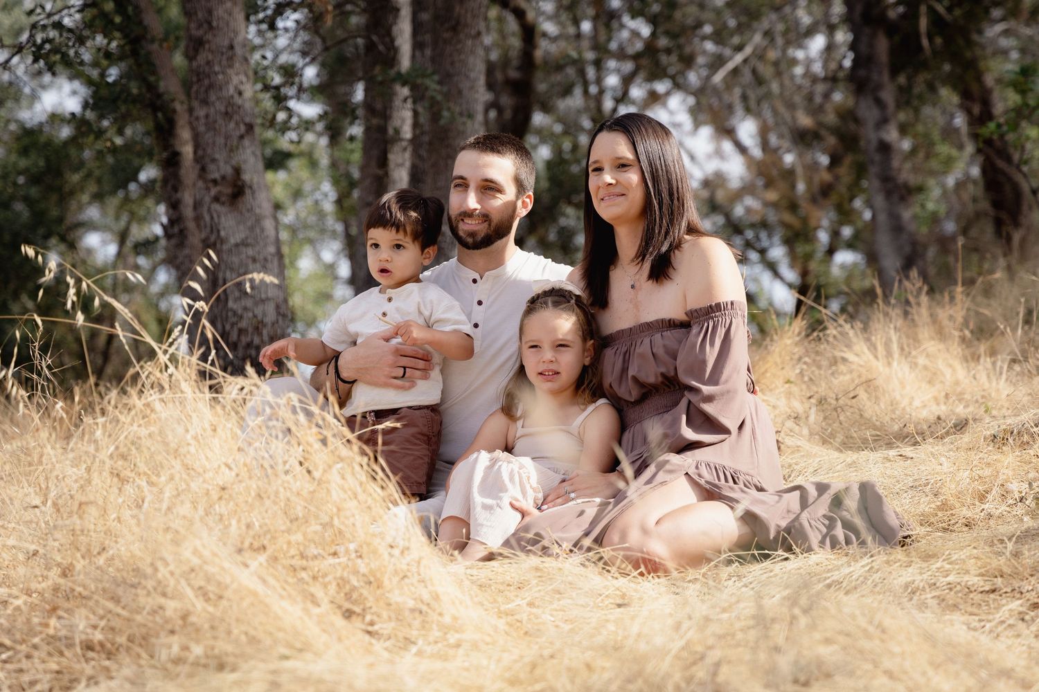 Family of four sitting in golden dry grass field among trees, parents with two young children in coordinating outfits.