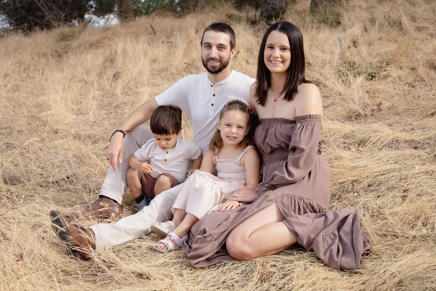 Family of four sitting together in a golden field, parents smiling with two young children in matching neutral outfits.