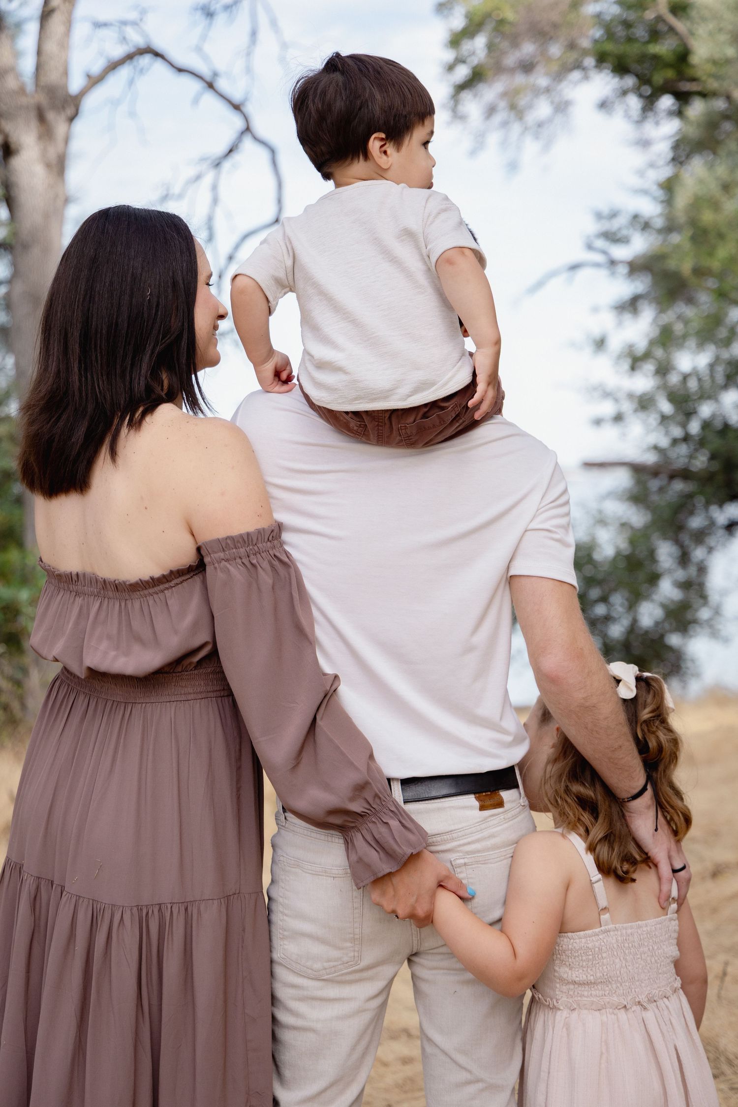 Family of four viewed from behind, parents holding hands with young children outdoors among trees.