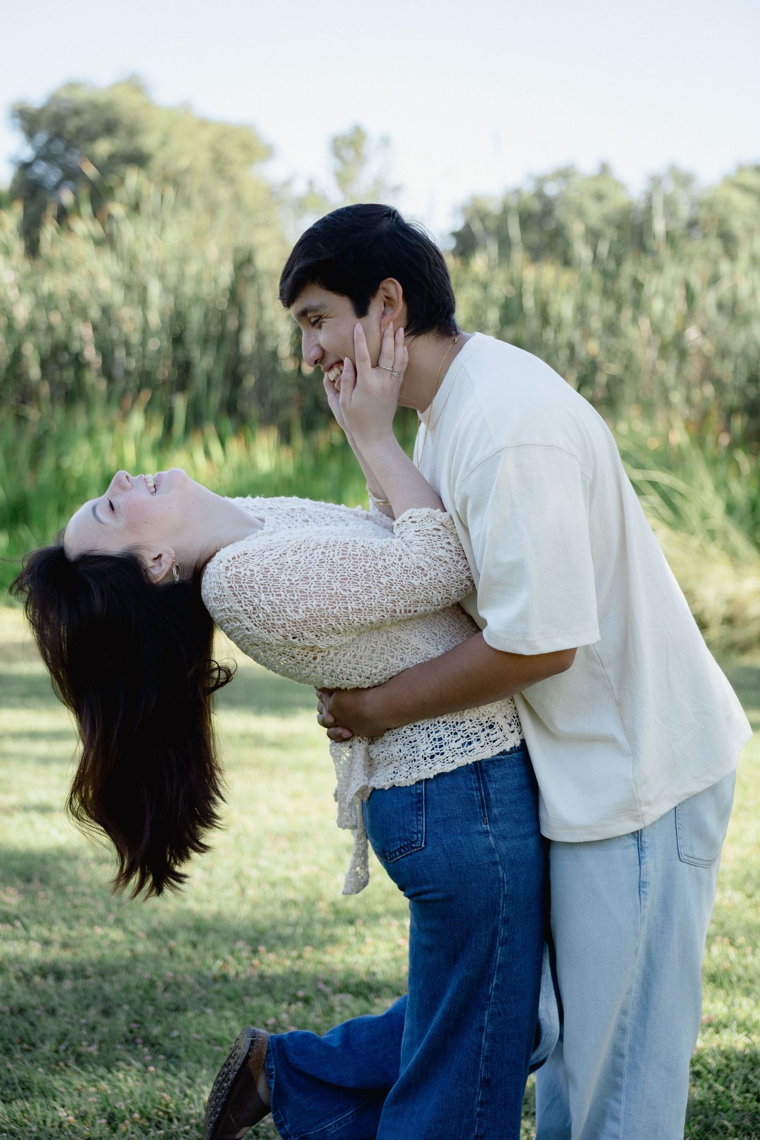 A joyful couple embraces outdoors, with the woman leaning back laughing and the man smiling in a lush green park.