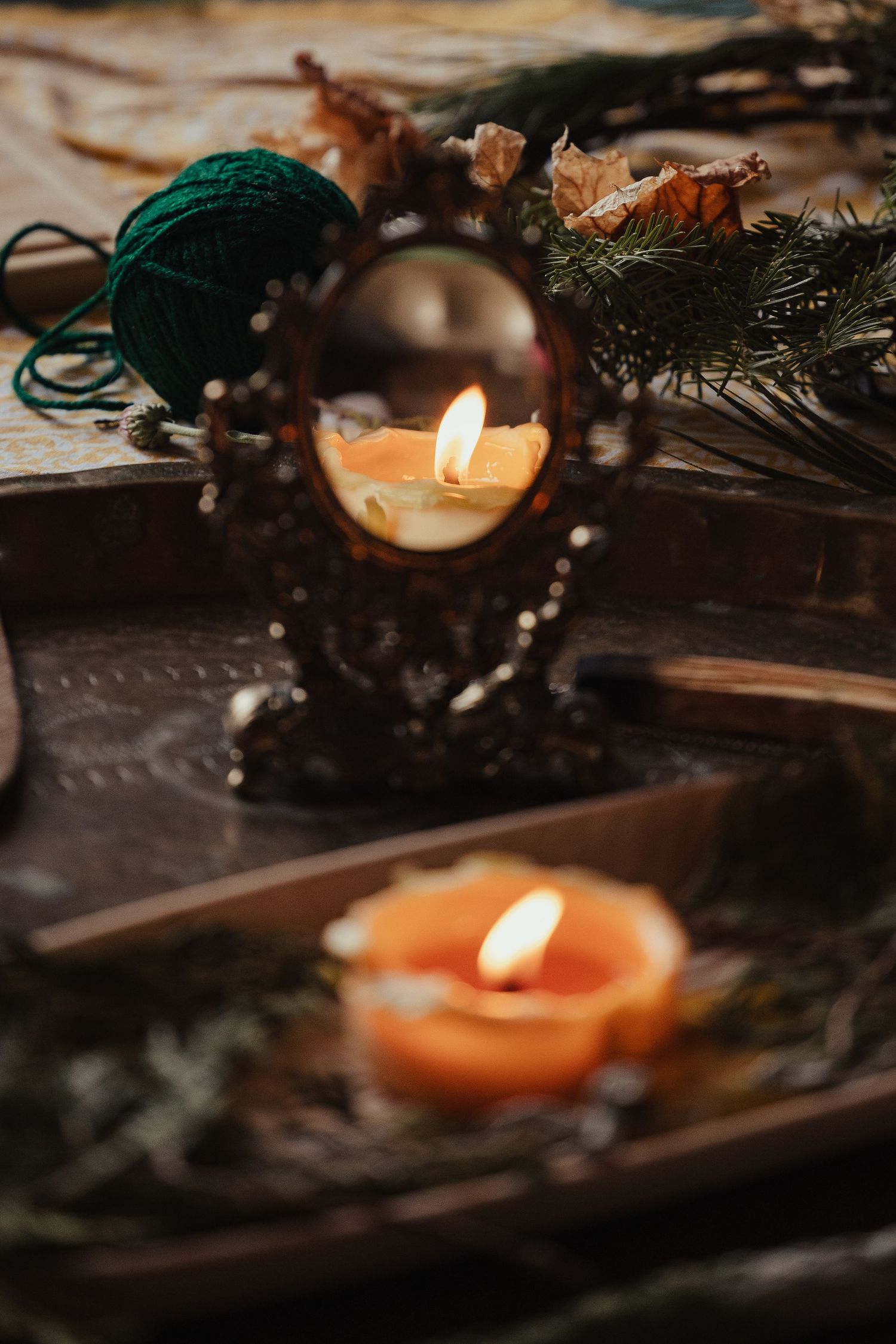 Mystical altar with lit orange candles, ornate mirror, green yarn, and pine branches on a rustic wooden tray.