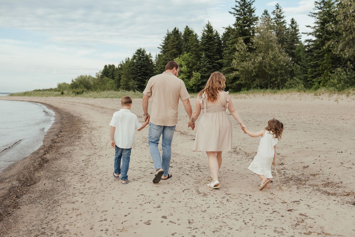 Family of four walking hand-in-hand along a sandy beach with lush green trees in the background.