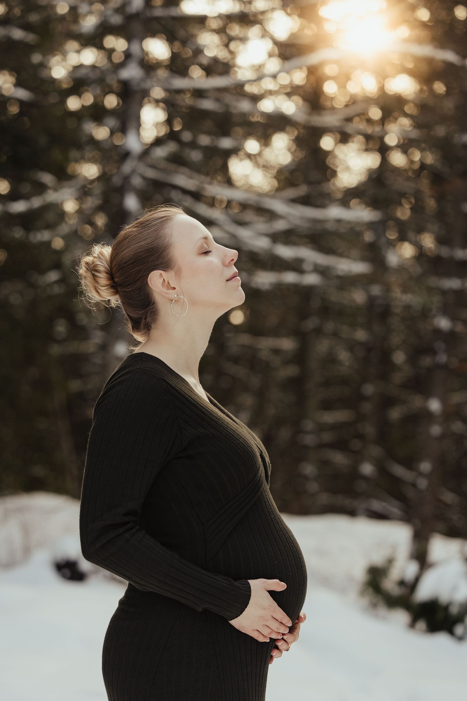 Pregnant woman in black dress holding belly outdoors in snowy winter forest with golden sunset light.