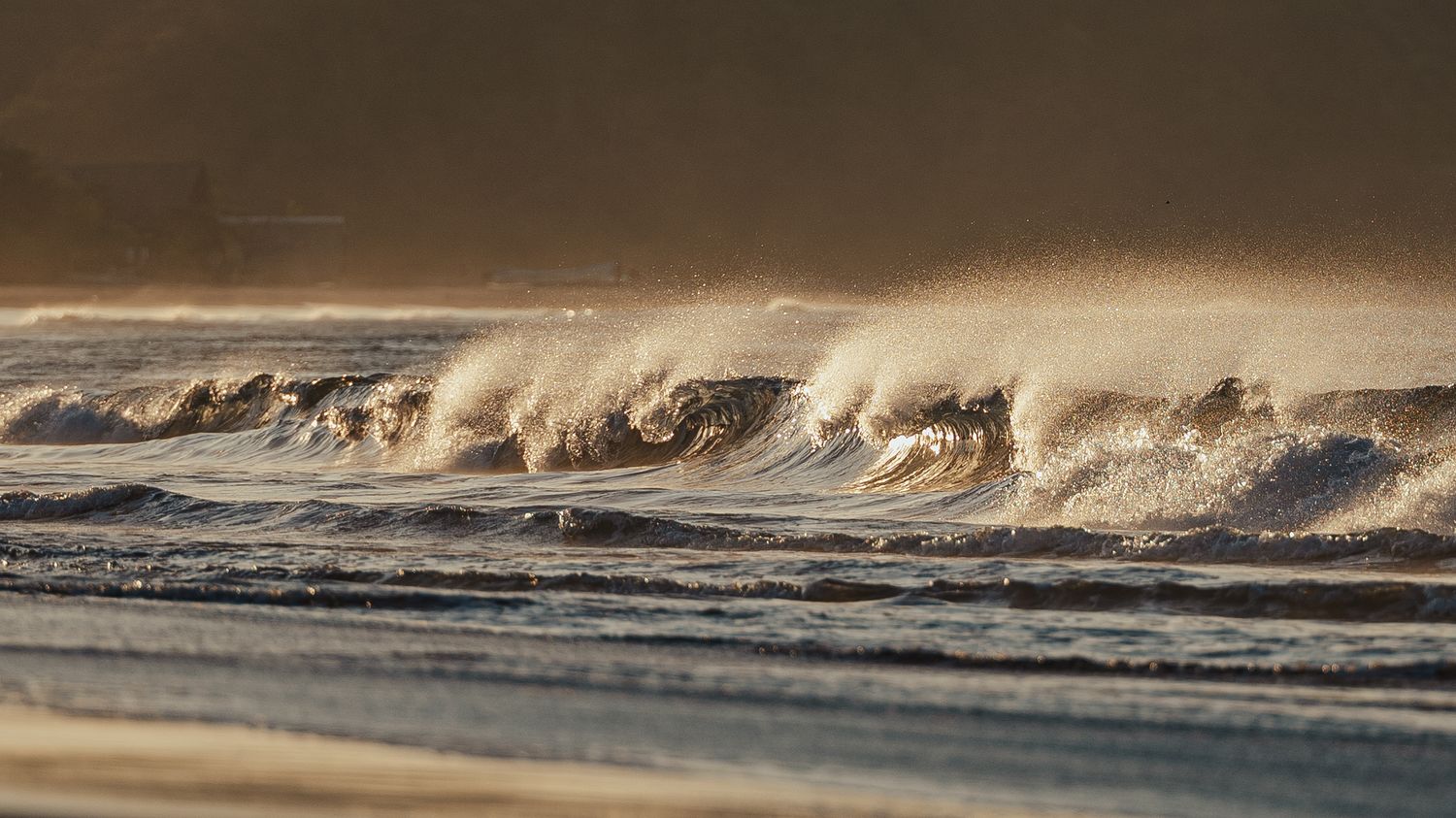 Powerful ocean waves crashing on a beach at golden hour, with sea spray and mist illuminated by warm sunlight.