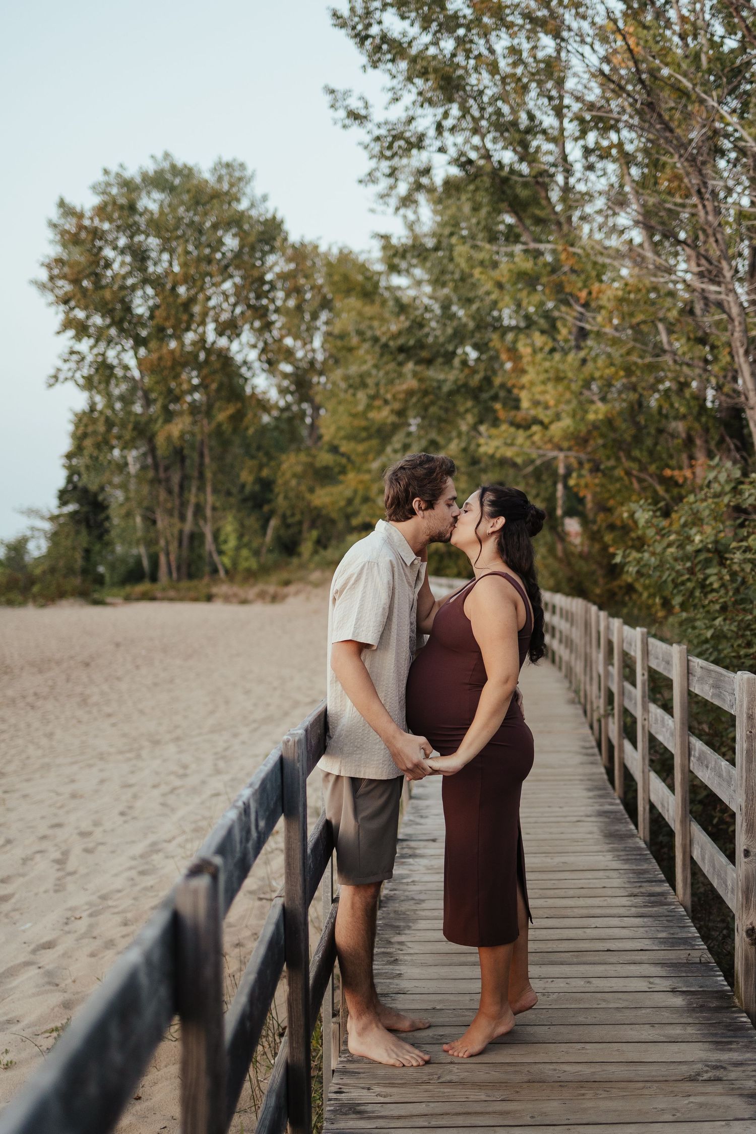 Pregnant couple kissing on a wooden boardwalk near a sandy beach surrounded by lush green trees.