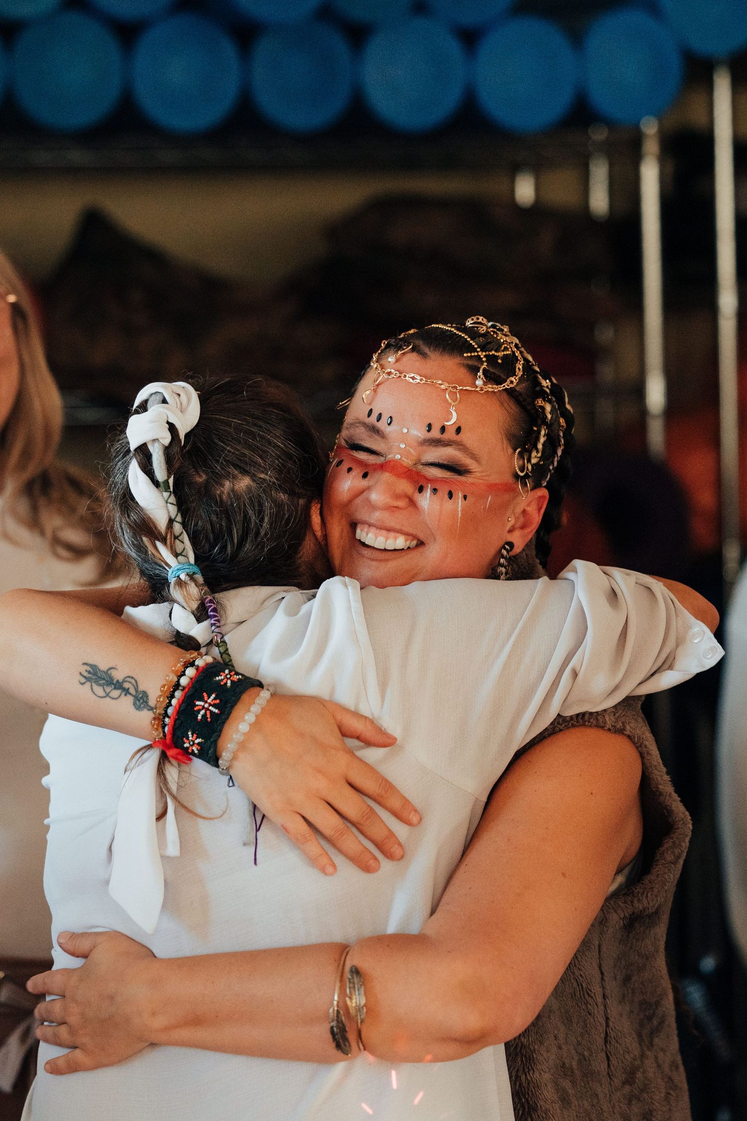 Two women sharing a joyful hug, one with decorative face paint and gold hair accessories, wearing bracelets at an outdoor event.
