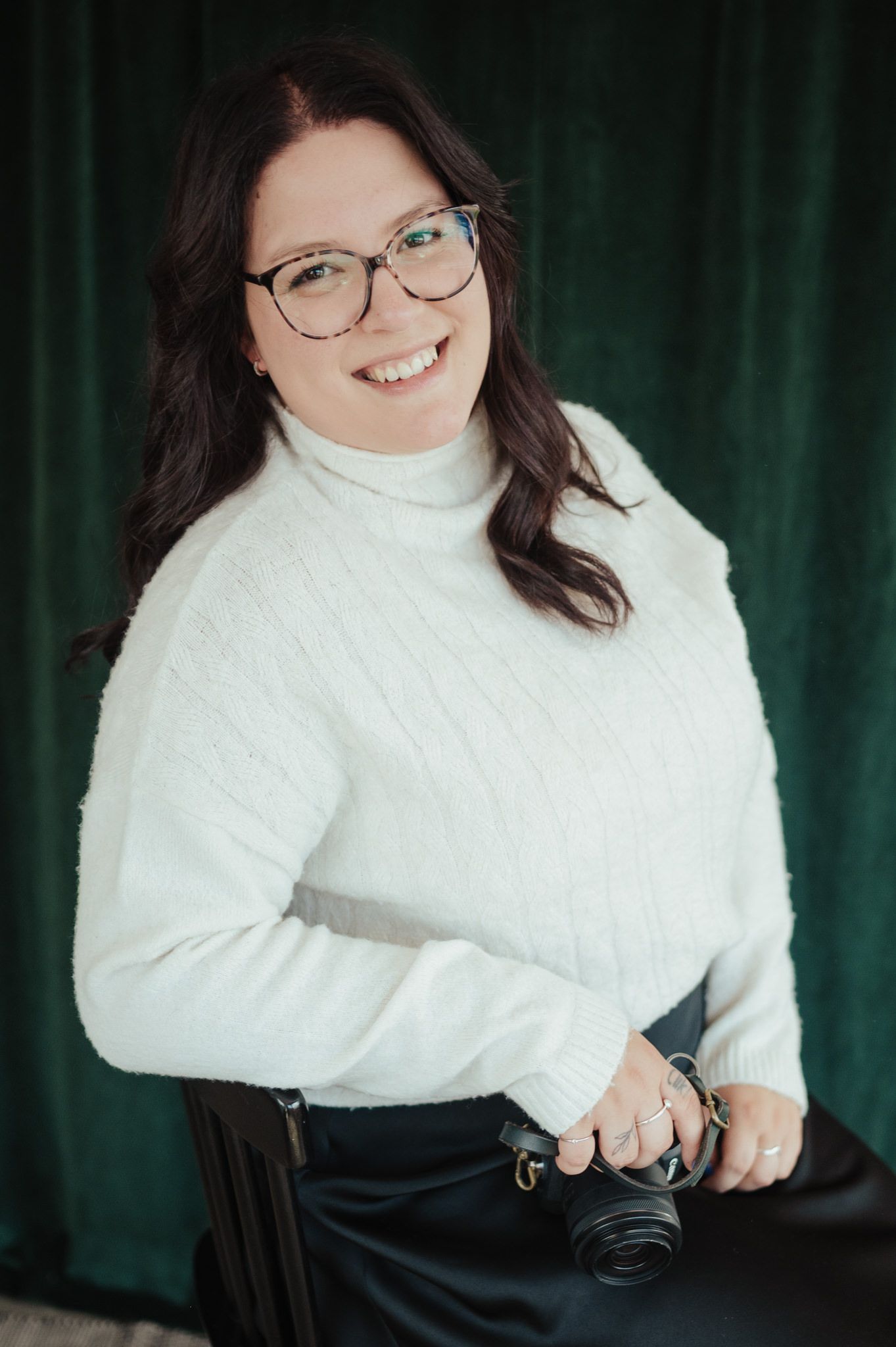 Smiling woman with dark hair and glasses wearing a white turtleneck sweater holding a camera against green backdrop.