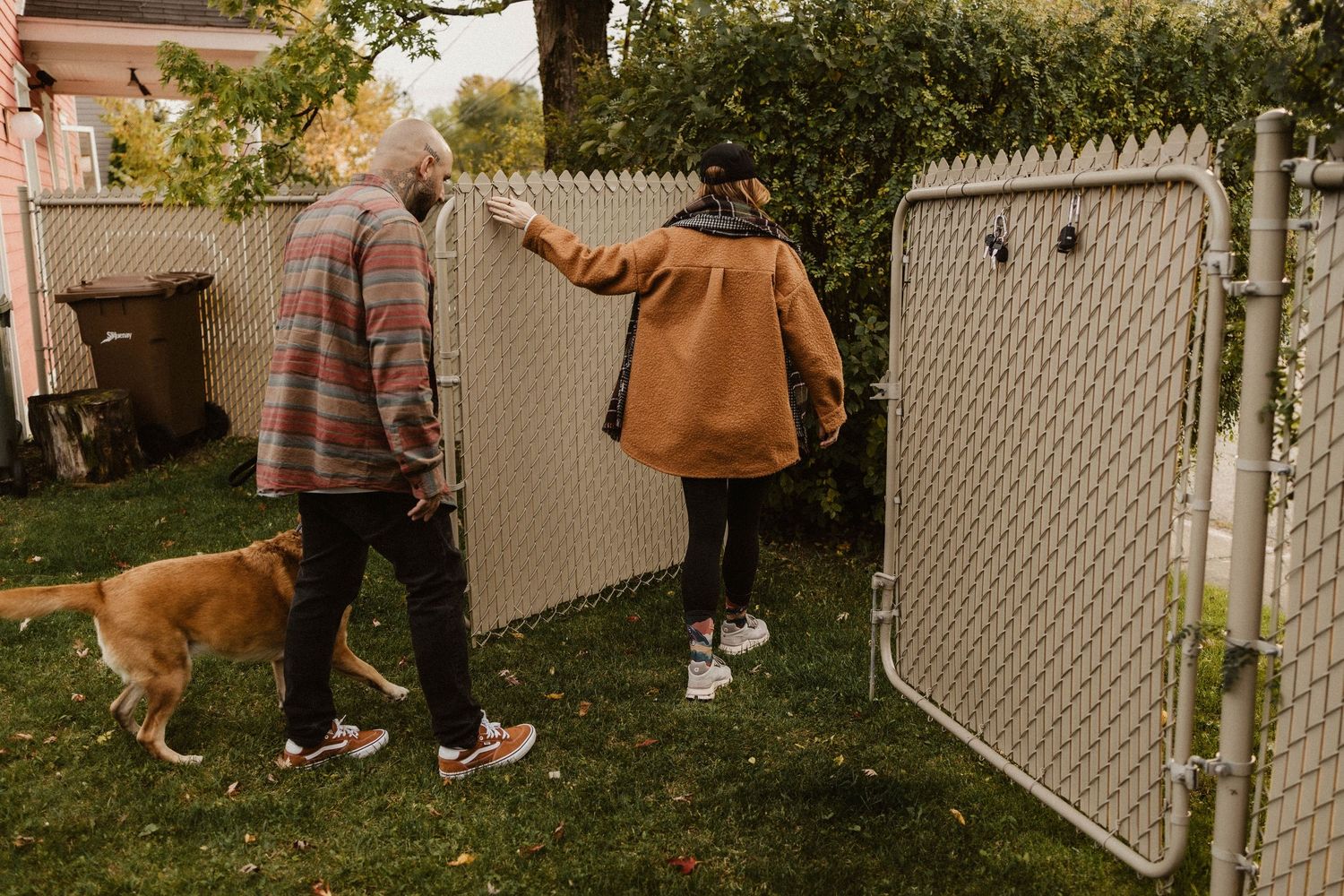Two people and a golden dog walk through a backyard gate in a fenced yard on an autumn day.