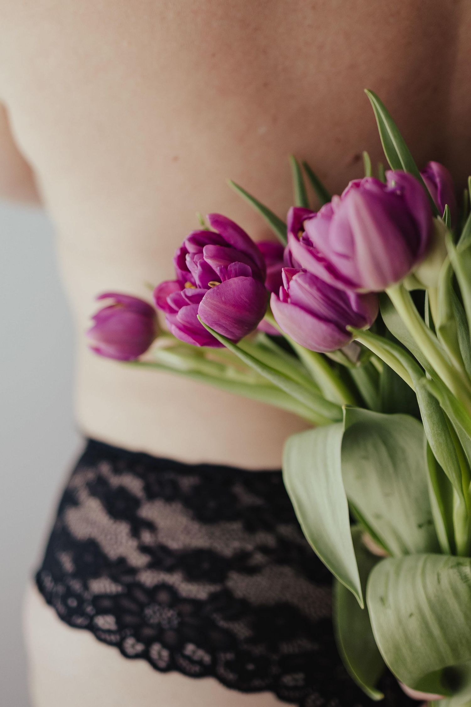 Woman in black lace underwear holding a bouquet of purple tulips behind her back.