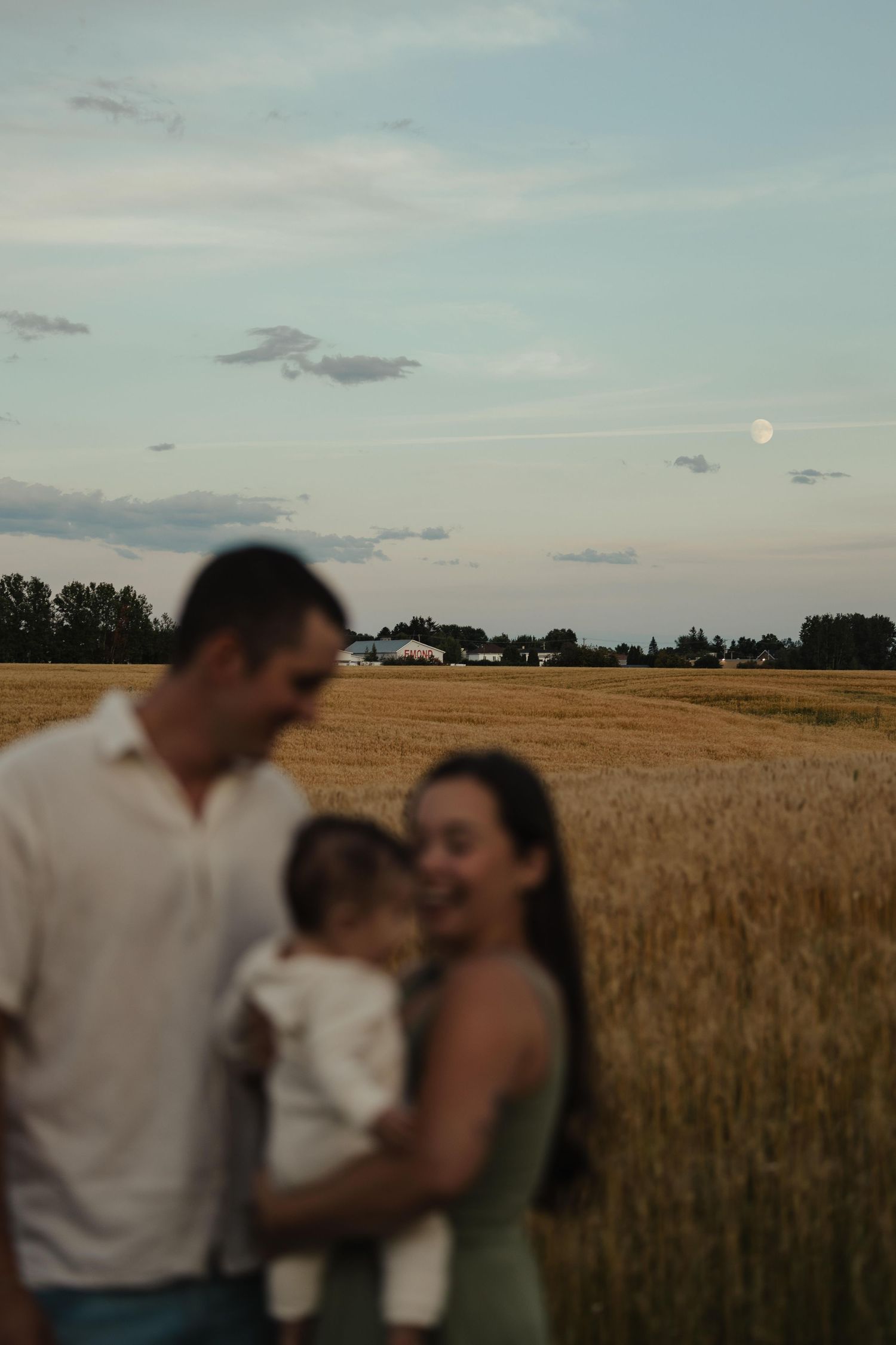 Family with baby standing in golden wheat field at dusk, full moon visible in pastel sky above rural farmland.