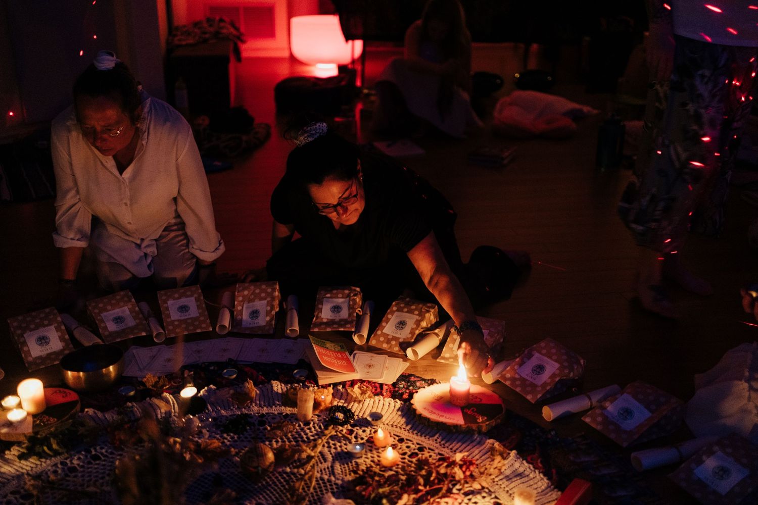 Two women arranging tarot cards and candles on floor in dimly lit room during a spiritual ritual ceremony.