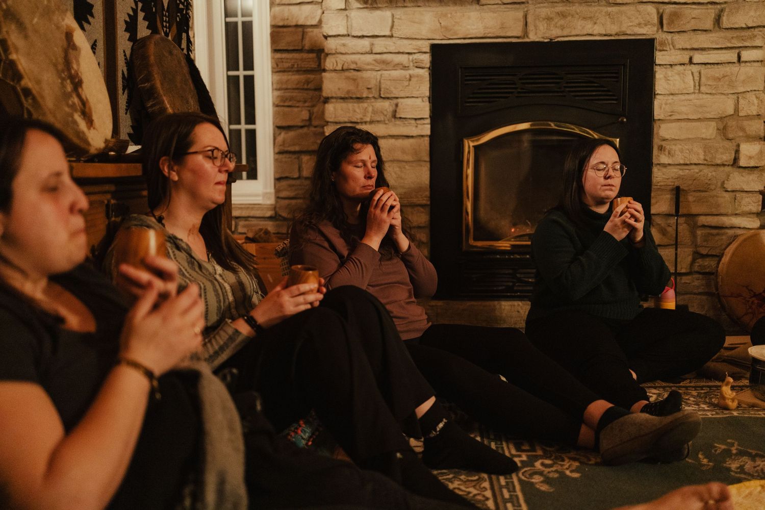 Women sitting by a cozy stone fireplace holding drinks in a warm intimate gathering.