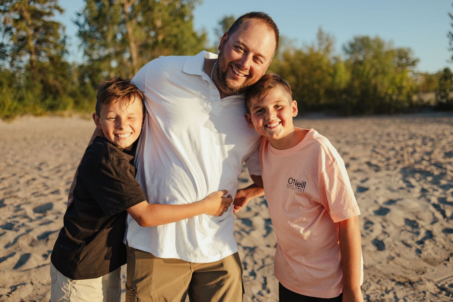 A smiling man hugs two children on a sandy beach, posing together for a warm family photo outdoors.