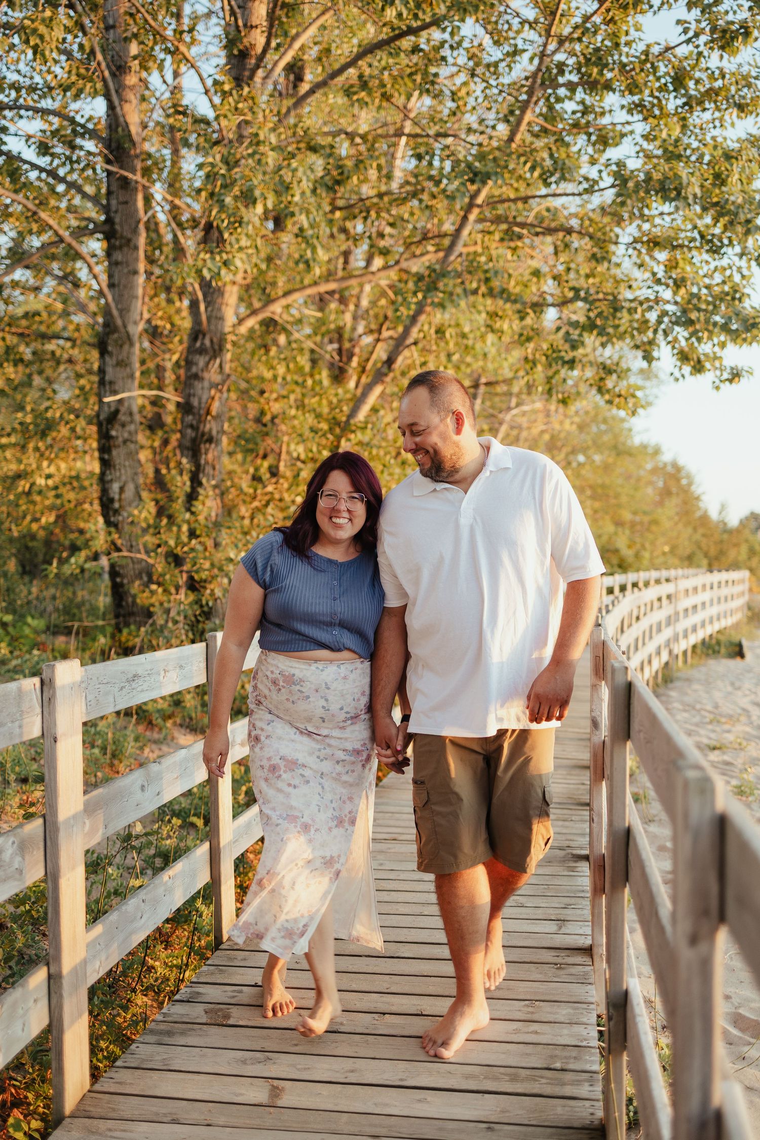 Happy couple walking barefoot on a wooden boardwalk surrounded by autumn trees in golden sunlight.