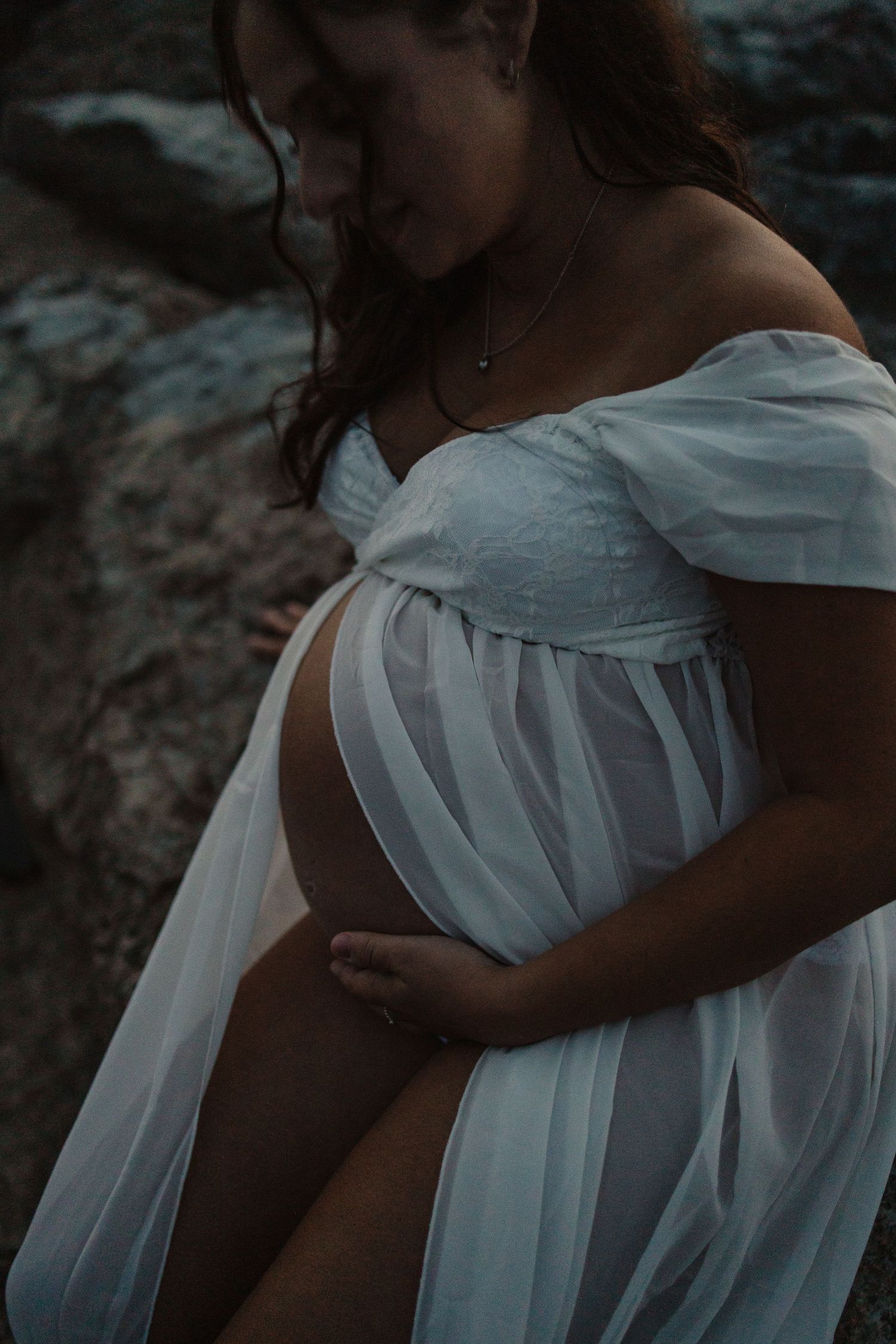 Pregnant woman in sheer white off-shoulder maternity gown cradling her baby bump outdoors near rocks.