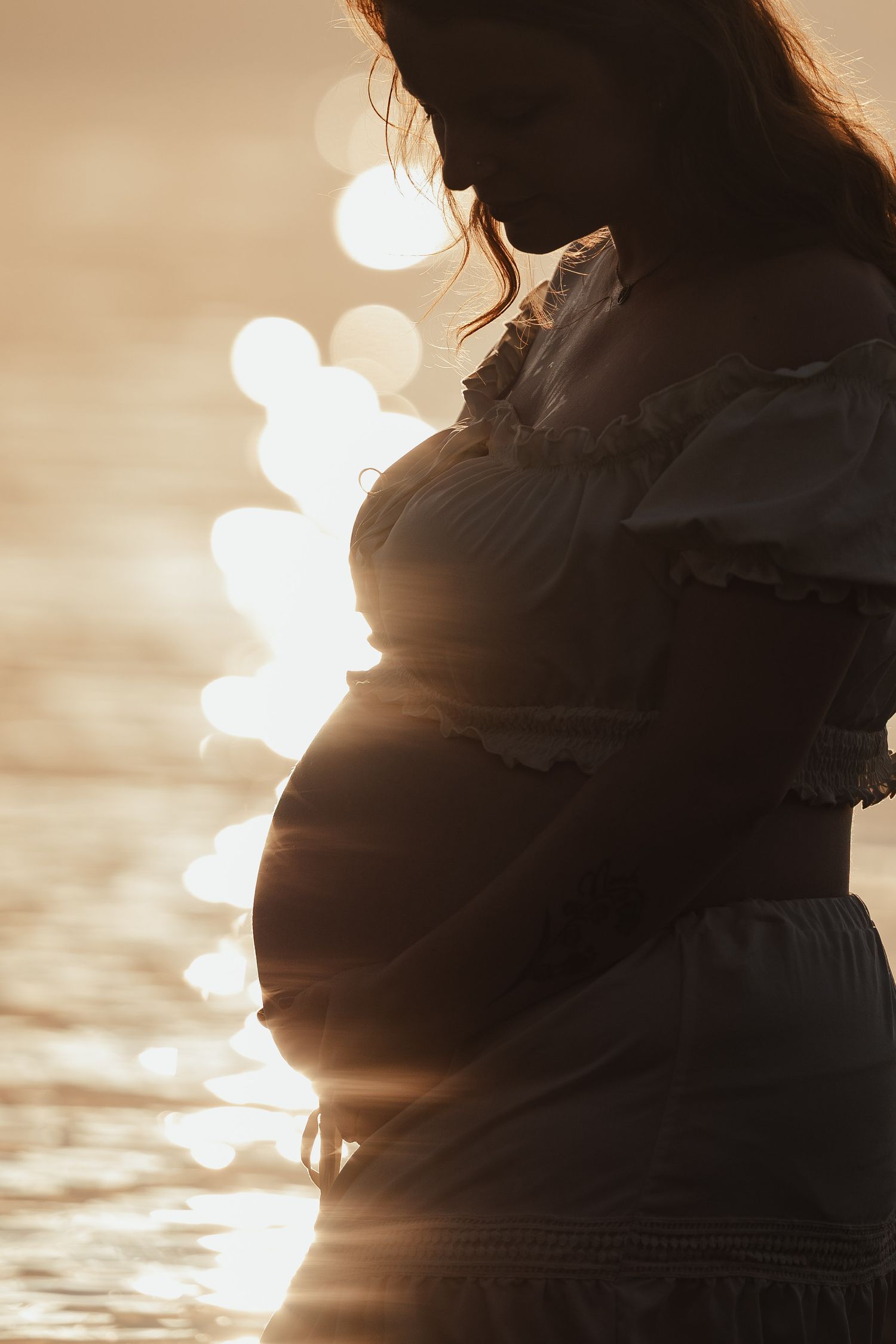 Silhouette of a pregnant woman in a white dress glowing in warm golden sunset light.