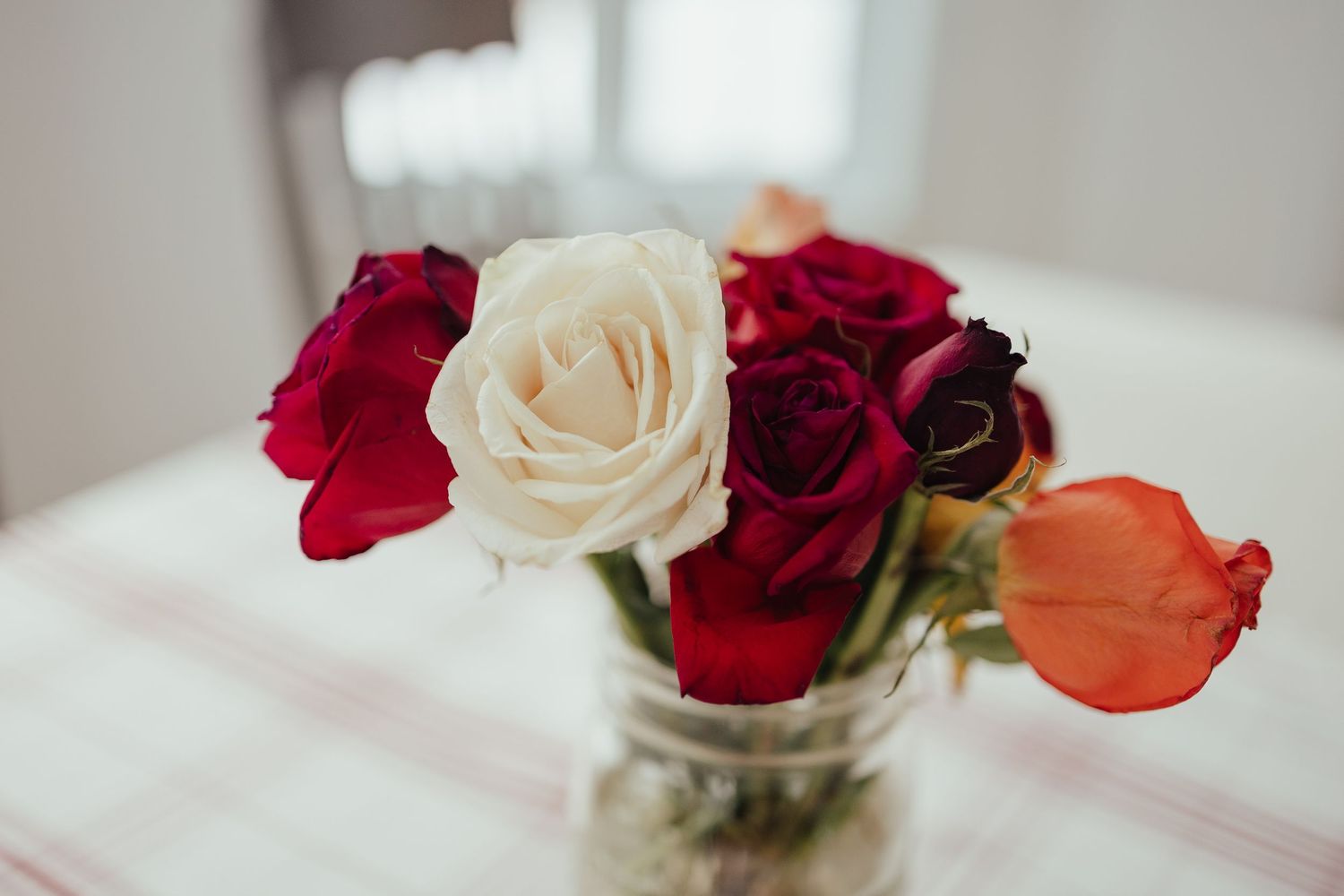 A glass jar vase with red, white, and orange roses arranged on a white striped tablecloth.