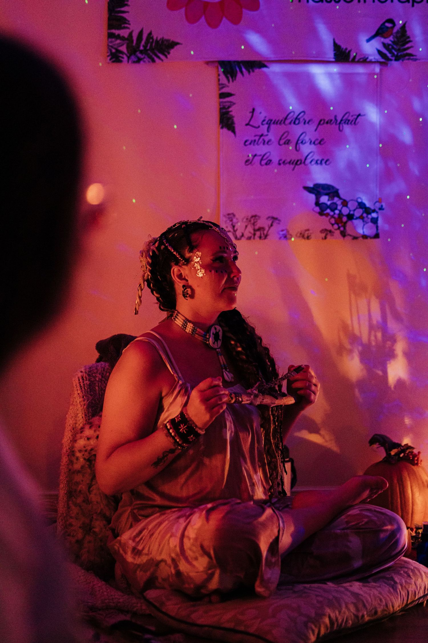 Woman sitting cross-legged in purple-lit room with French text projection, wearing jewelry and bohemian attire.