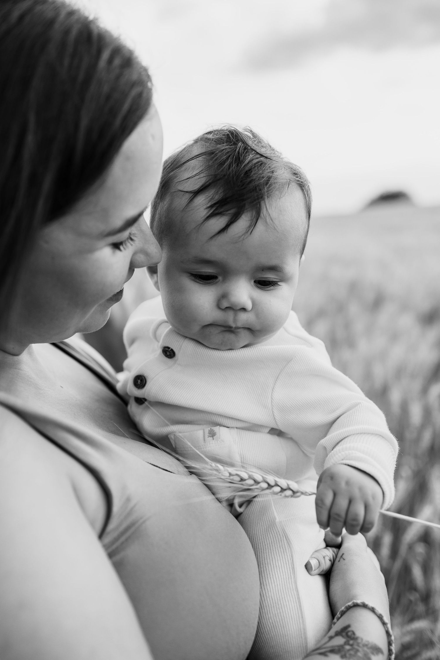 Black and white photo of a mother holding her baby in a wheat field, the infant grasping a wheat stalk.