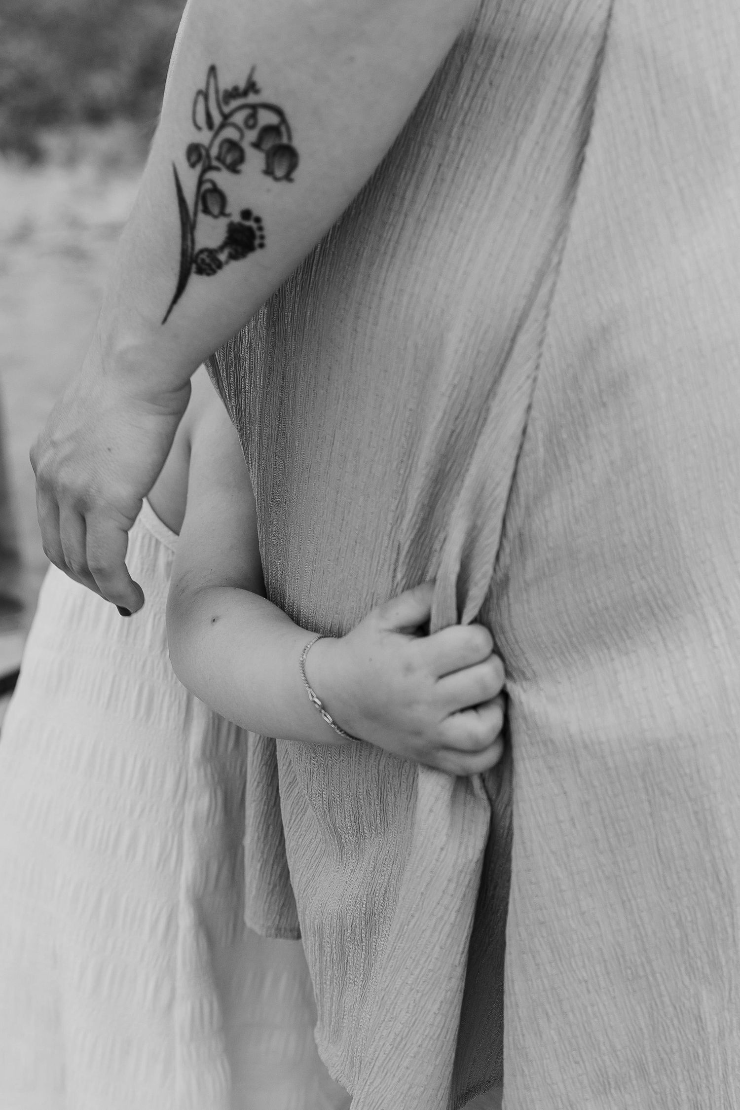 Black and white photo of a mother with a floral tattoo holding her baby's hand, child gripping her dress.