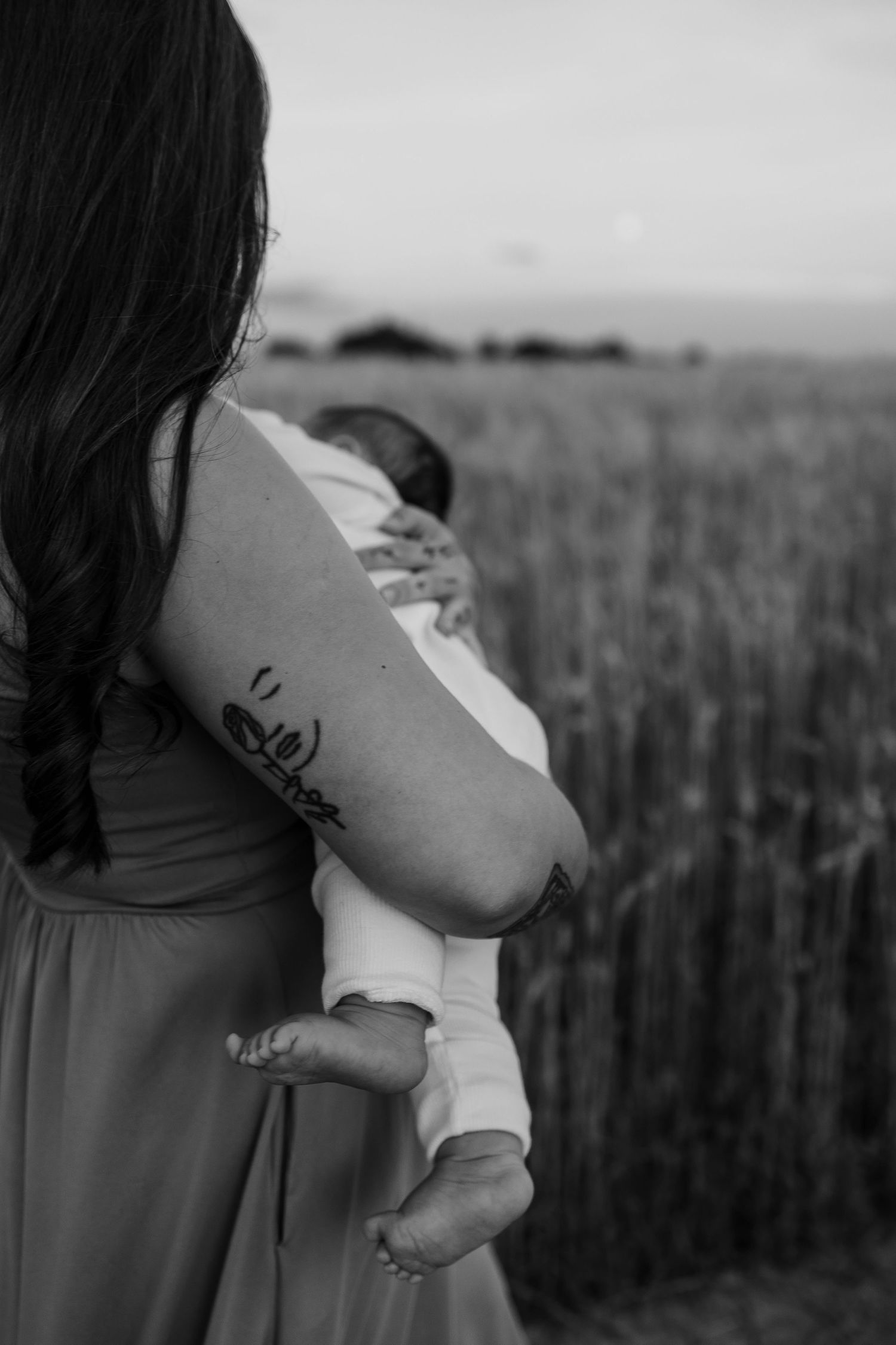 Black and white photo of mother with floral arm tattoo cradling newborn baby in wheat field.