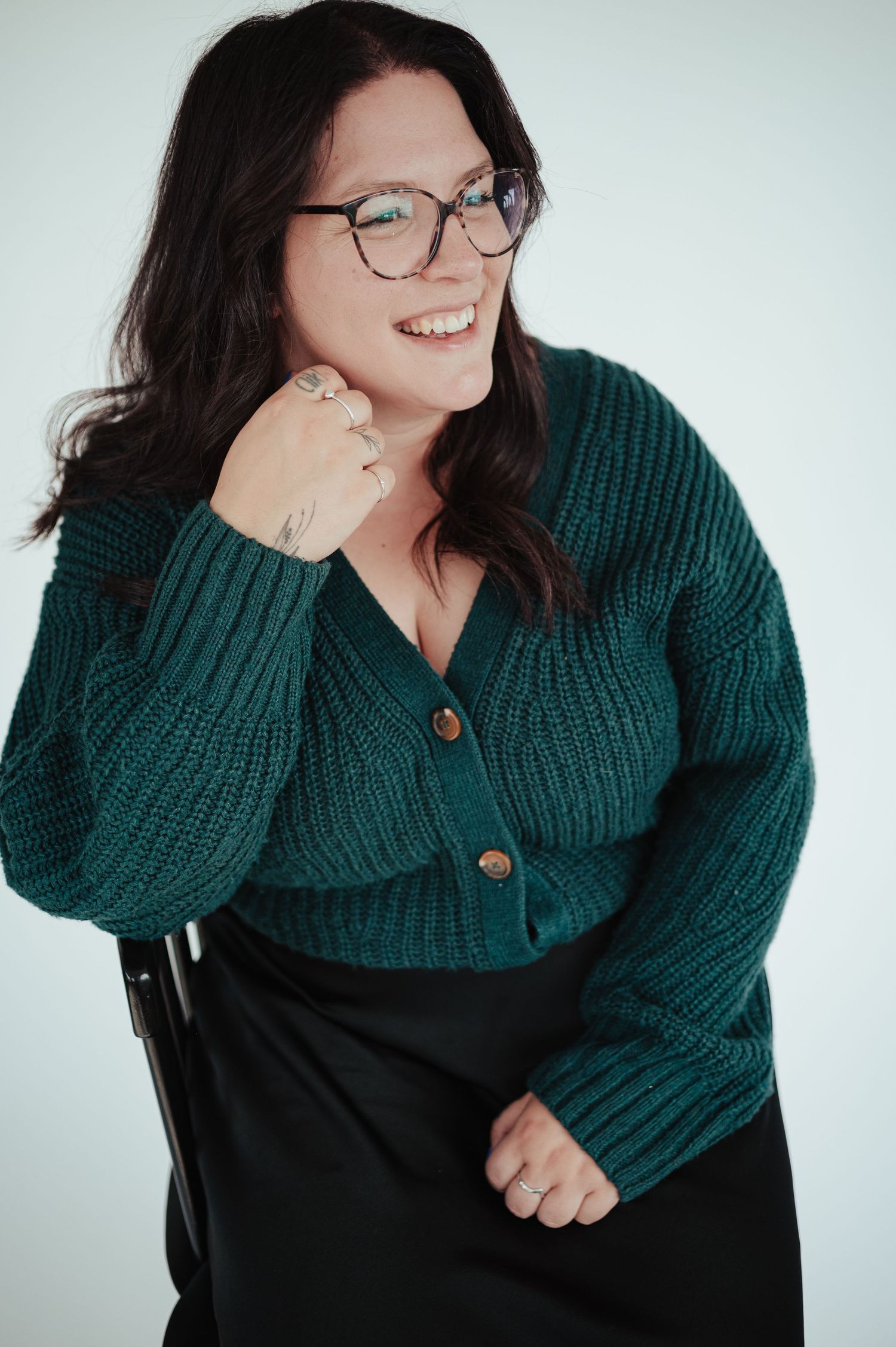 Smiling woman with glasses wearing a teal knit cardigan and black skirt poses confidently against a light background.