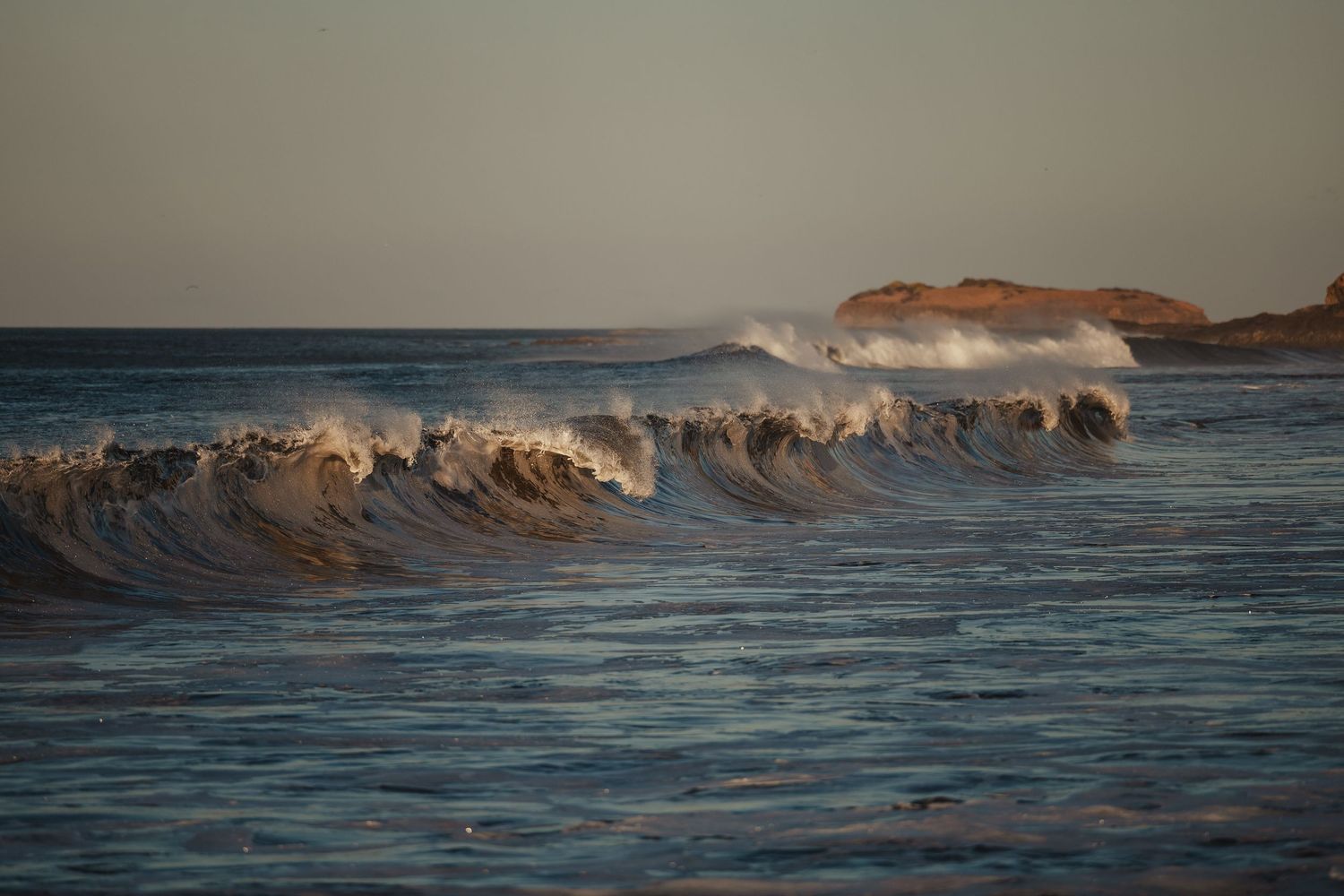 Ocean waves crashing near rocky coastline at golden hour sunset light.