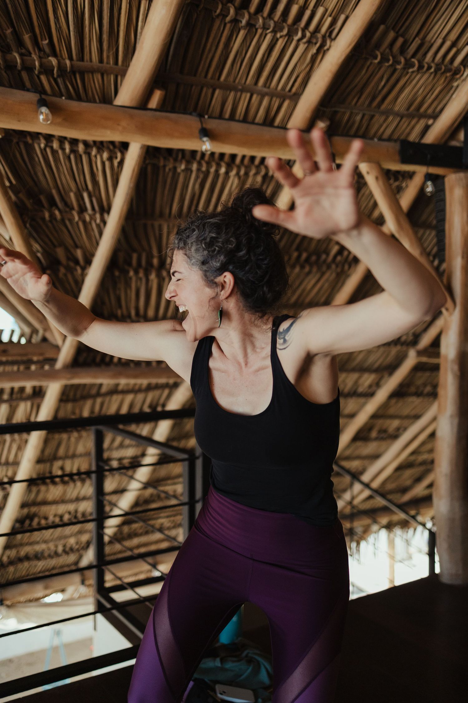 Woman in black tank top and purple leggings practicing yoga under a rustic bamboo thatched roof.