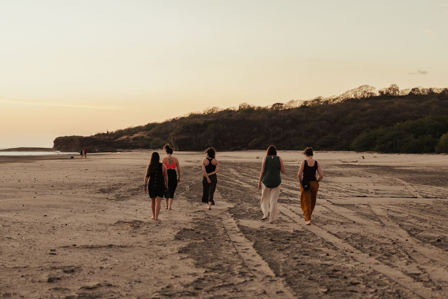 Four women walking along a sandy beach at sunset with tree-covered hills in the background.