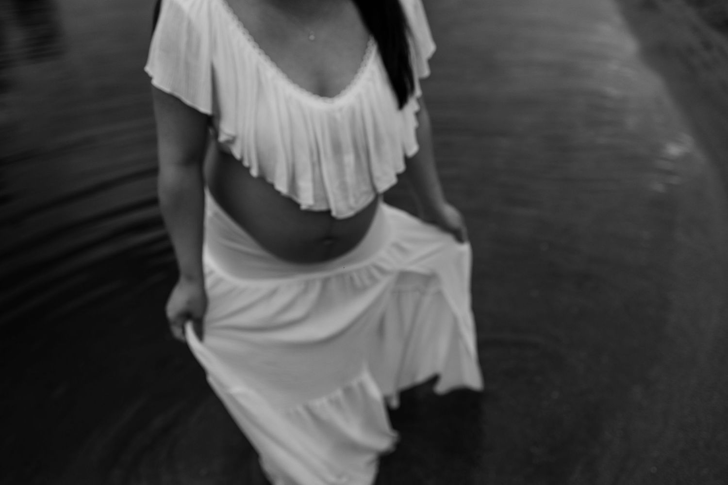 Black and white photo of a pregnant woman in a white crop top and maxi skirt standing in water.