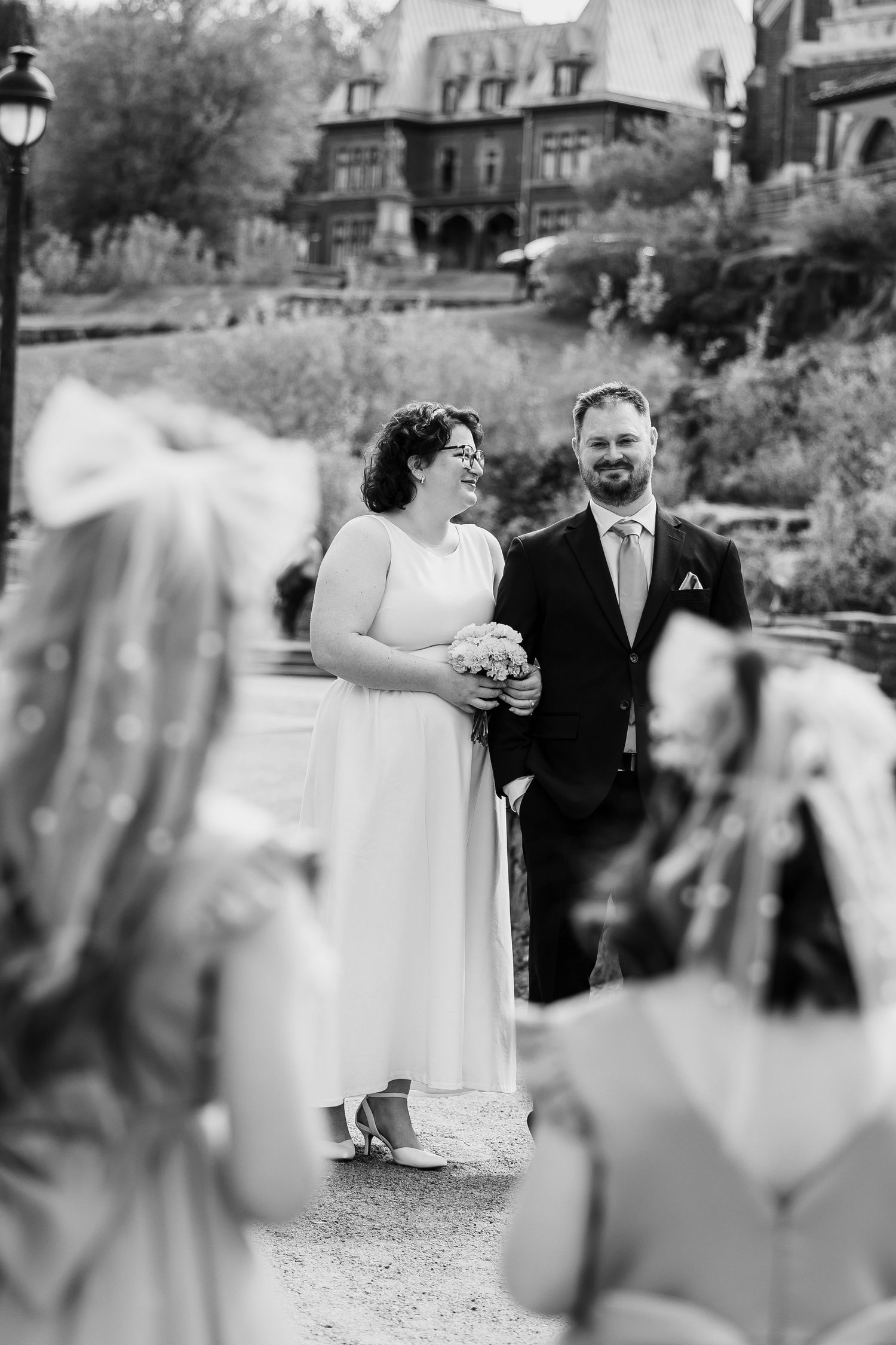 Black and white photo of a smiling bride holding bouquet and groom in suit standing outdoors at elegant estate wedding.