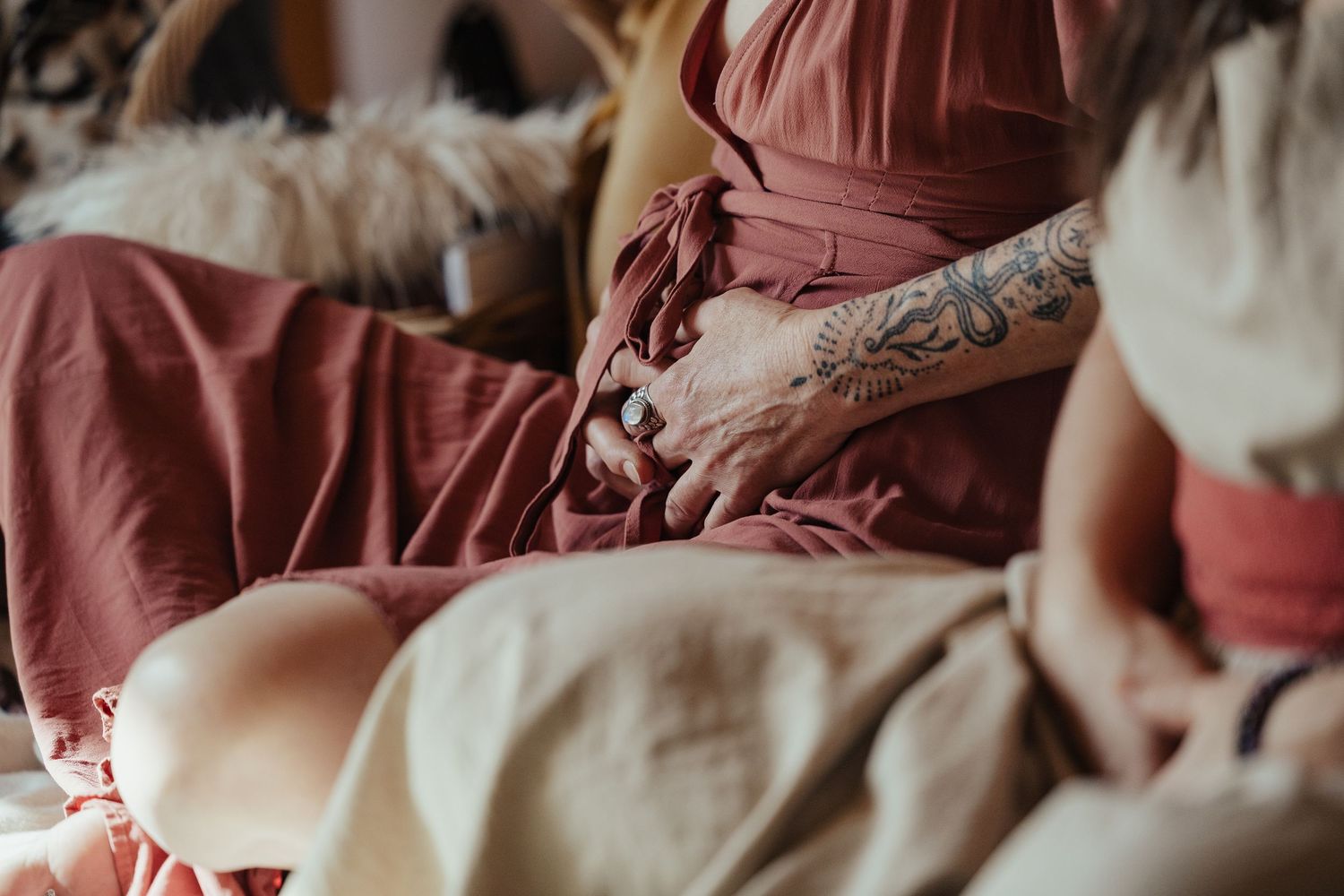 Pregnant woman in a rust-colored dress cradling her baby bump with a tattooed hand during maternity session.