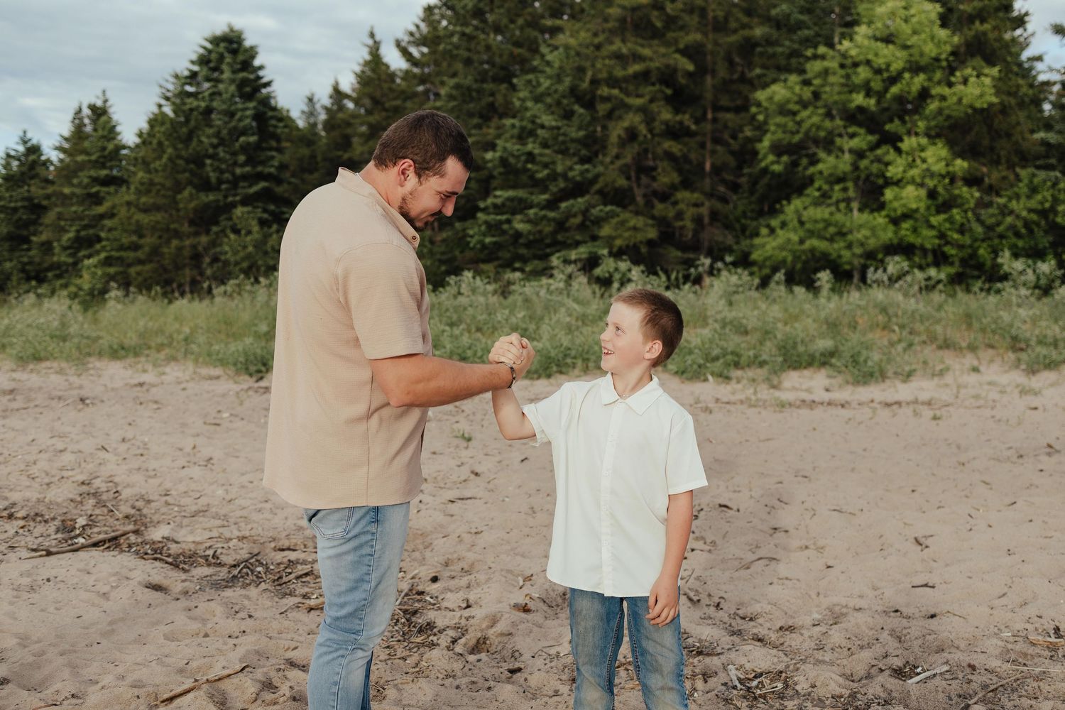 A father and young son share a joyful fist bump on a sandy beach with green trees in the background.