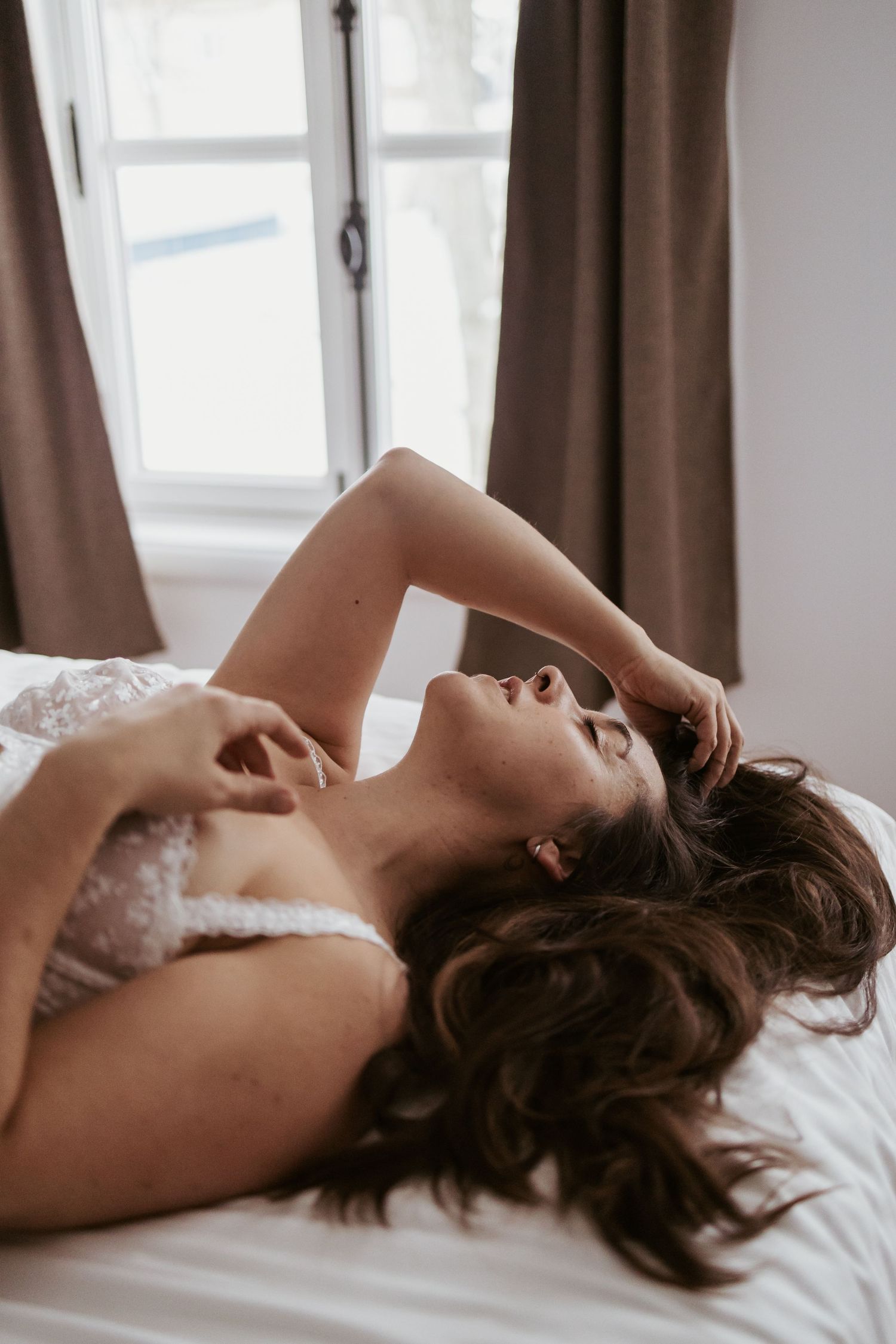 Woman in white lace lingerie lying on a bed with dark curtains and window in the background.