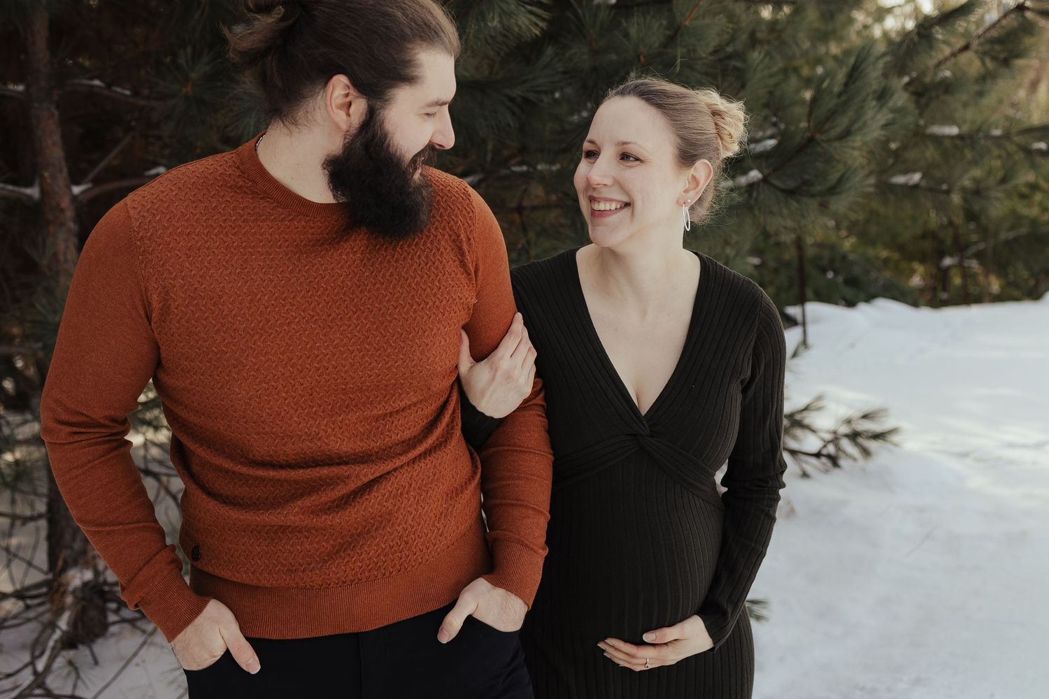Smiling couple in winter maternity photo, man in rust sweater and pregnant woman in black dress, snowy pine tree background.